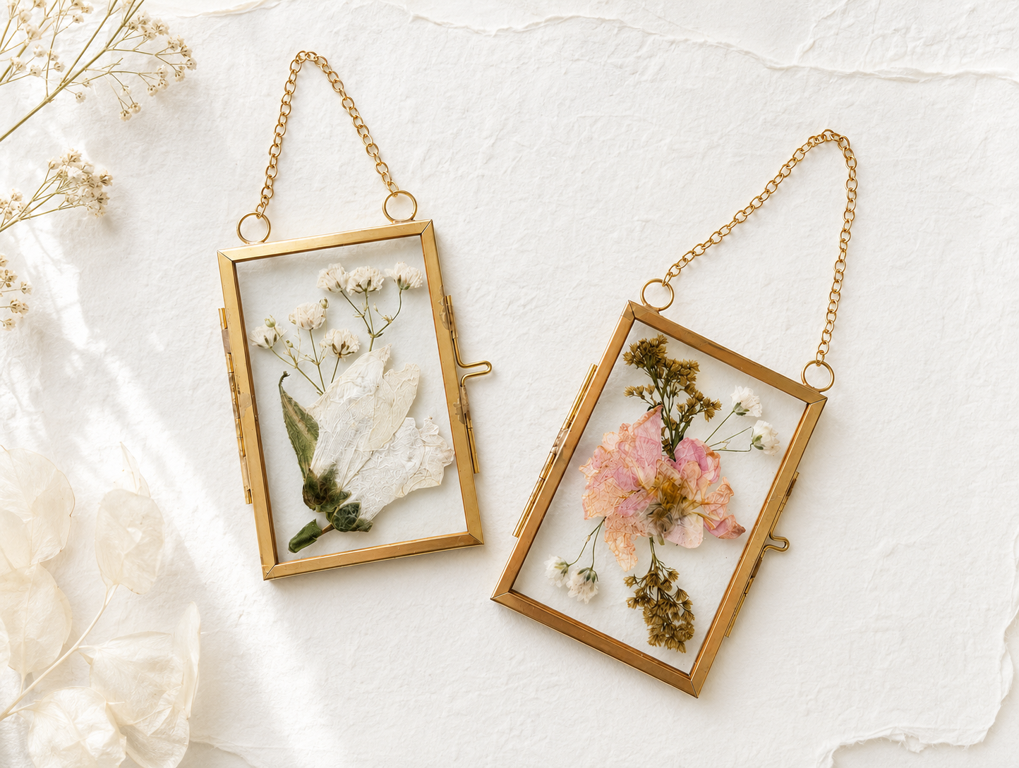Two gold rectangular pressed floral ornaments, each containing pressed flowers, placed on a white textured surface with additional dried flowers nearby.