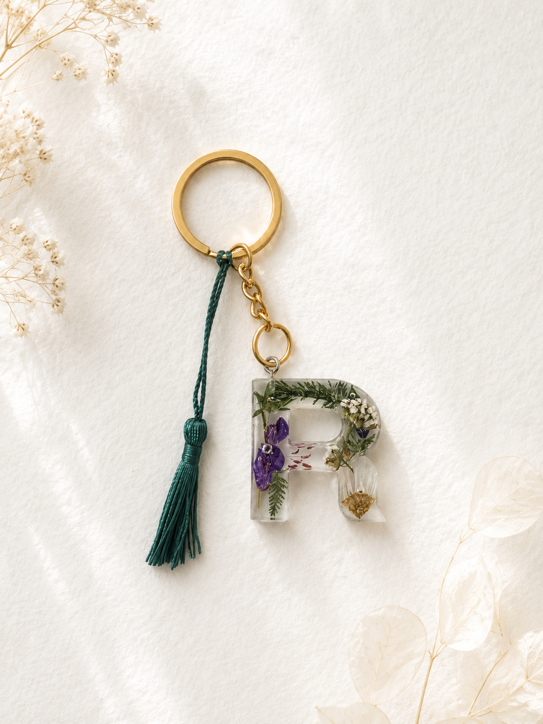 Resin keychain with a large gold ring, green tassel, and a decorative letter 'R' filled with dried flowers and greenery.