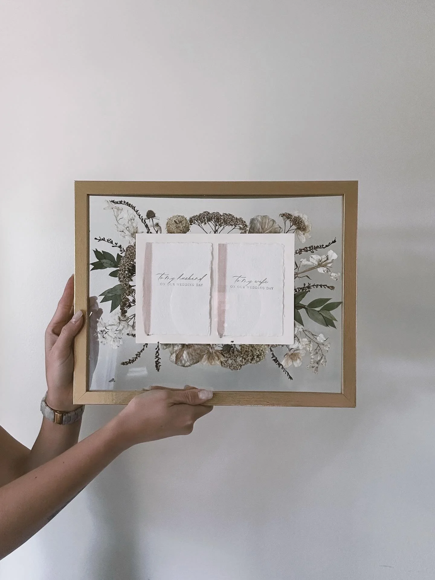 Person holding a framed artwork with dried flowers and two white cards that say "To my husband on our wedding day" and "To my wife on our wedding day" in front of a plain white wall.