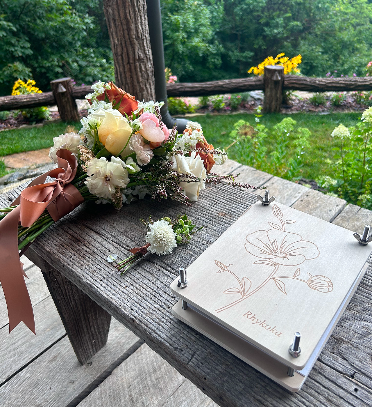 A bouquet of flowers with pink, white, and peach roses, white hydrangeas, and greenery, tied with a pink satin ribbon, resting on a rustic wooden table. Next to the bouquet is a white art board with a sketch of a flower and the name "Rhyokoa." The setting appears to be outdoors with a lush green garden and wooden fence in the background.