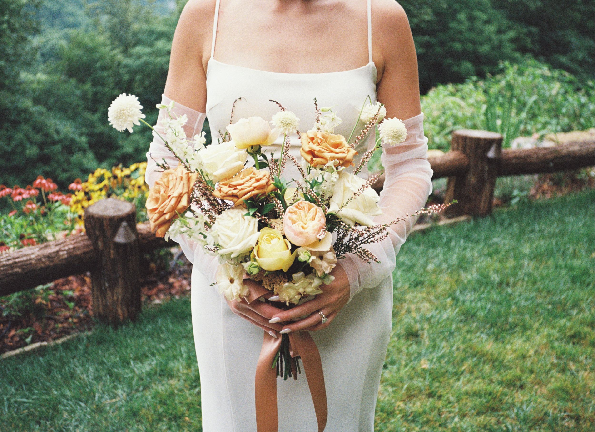 A woman in a white dress holding a bouquet of white and orange flowers outdoors, with a wooden fence and greenery in the background.