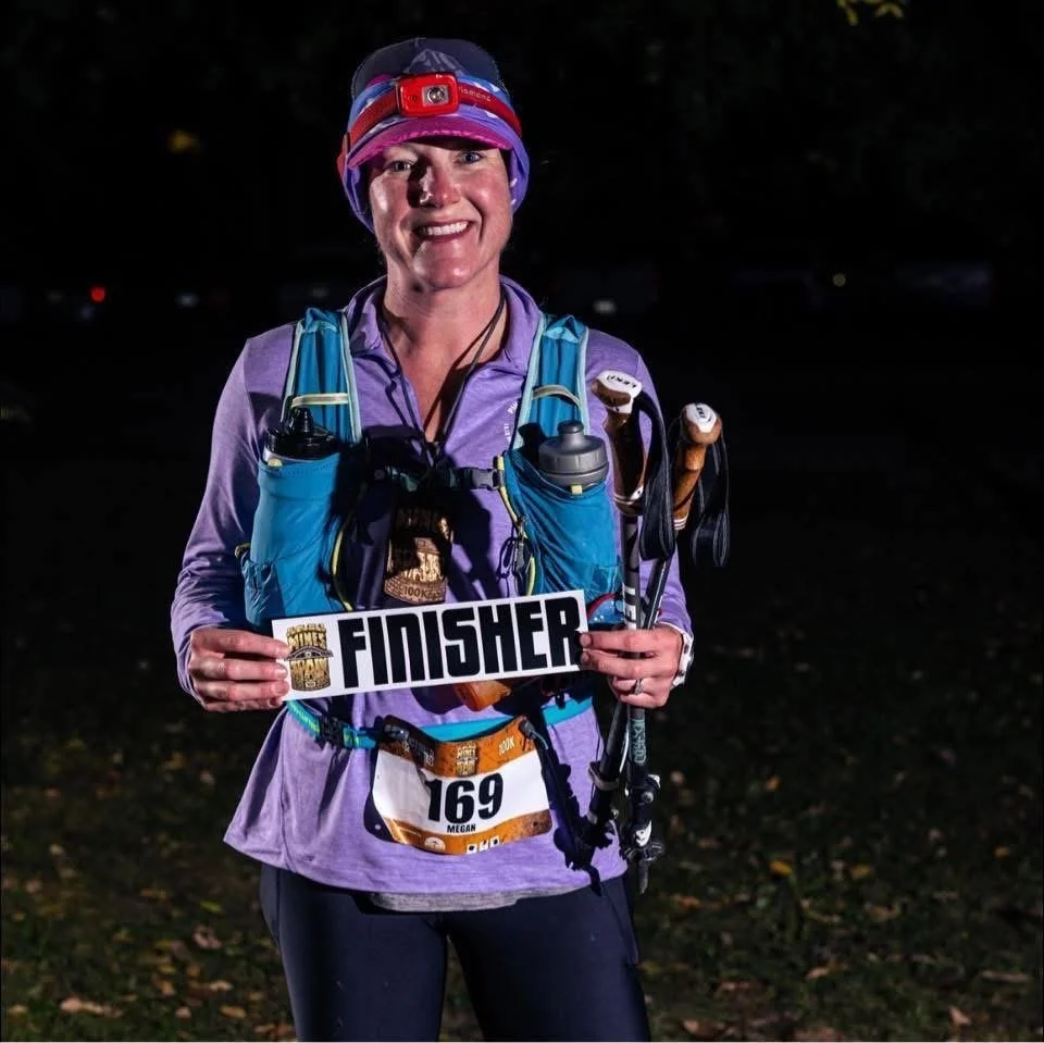 A woman in athletic gear holding a 'Finisher' sign and a race bib number 169, standing outdoors at night, smiling.