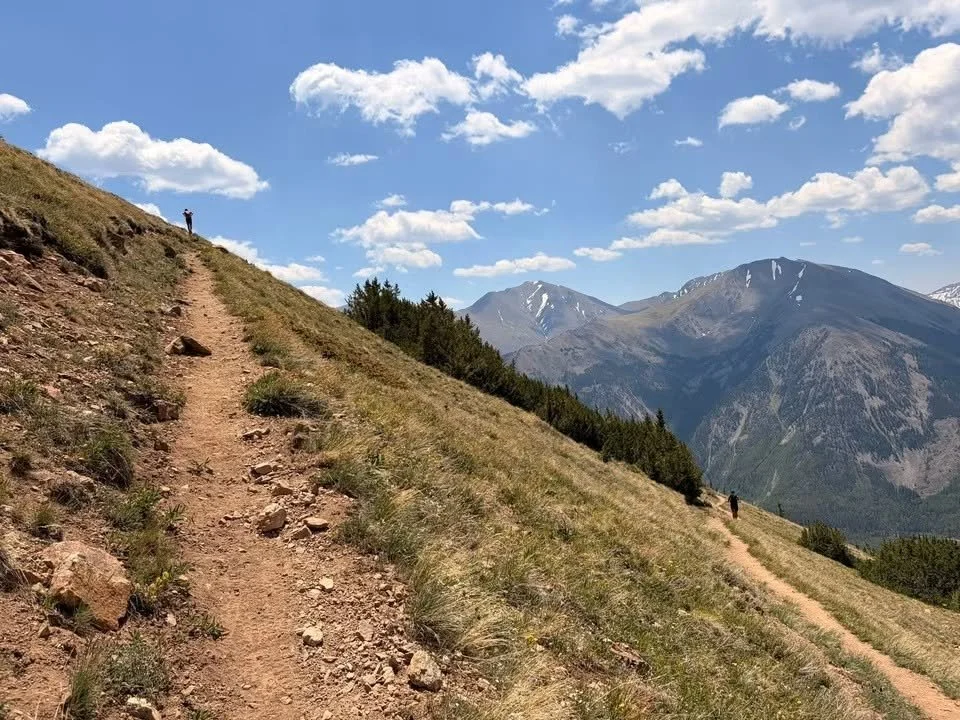Hiking trail on a grassy mountain slope with two hikers and mountain peaks in the background under a partly cloudy sky.