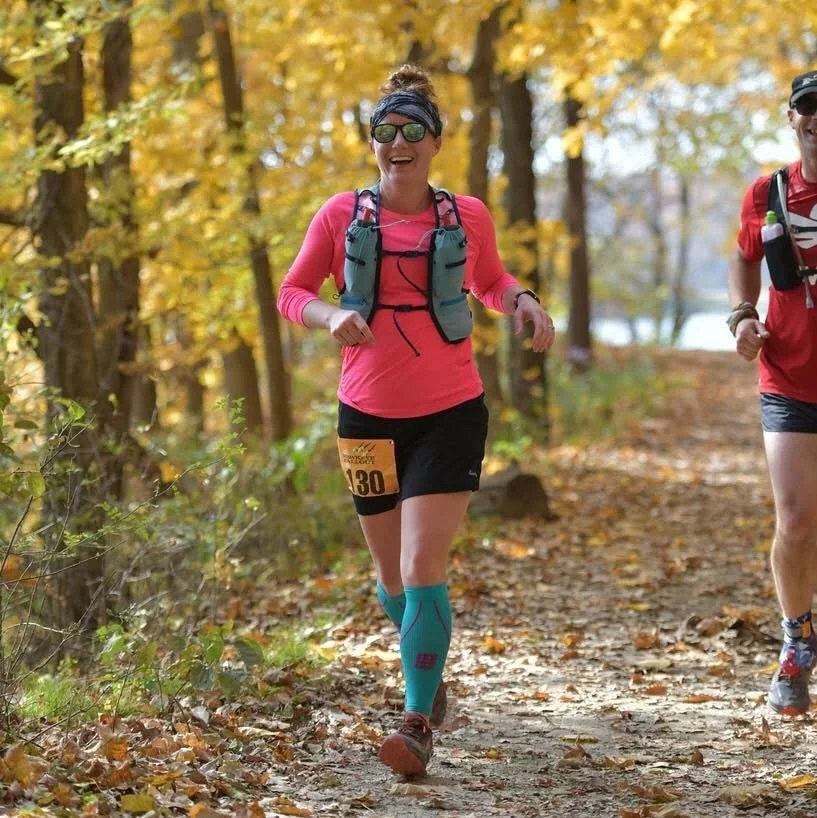 A woman smiling and running in a forest trail during autumn, wearing sunglasses, a pink long-sleeve shirt, black shorts, blue compression socks, and carrying a hydration vest.
