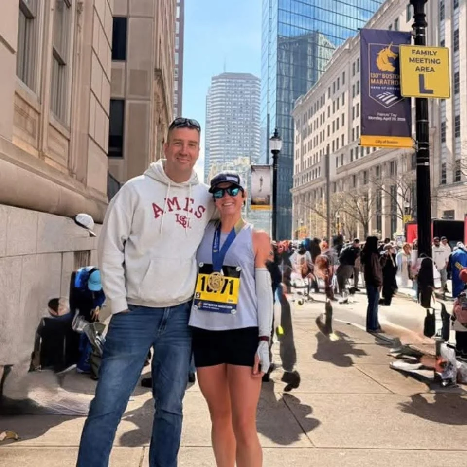 Two runners standing on a city sidewalk during a marathon, smiling at the camera. One is a man in a white hoodie and jeans, the other is a woman in running gear with a race bib and medal. Tall buildings and a crowd are in the background.