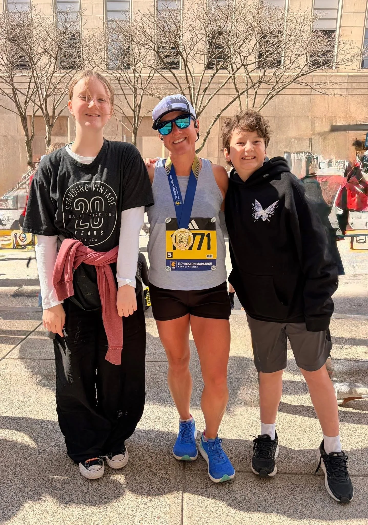 Three people standing together outdoors after a race, two children and an adult woman, smiling. The woman is wearing sunglasses, a cap, and a race medal, with a race bib number 1071 for the Boston Marathon. The children are wearing casual athletic clothing and sneakers.