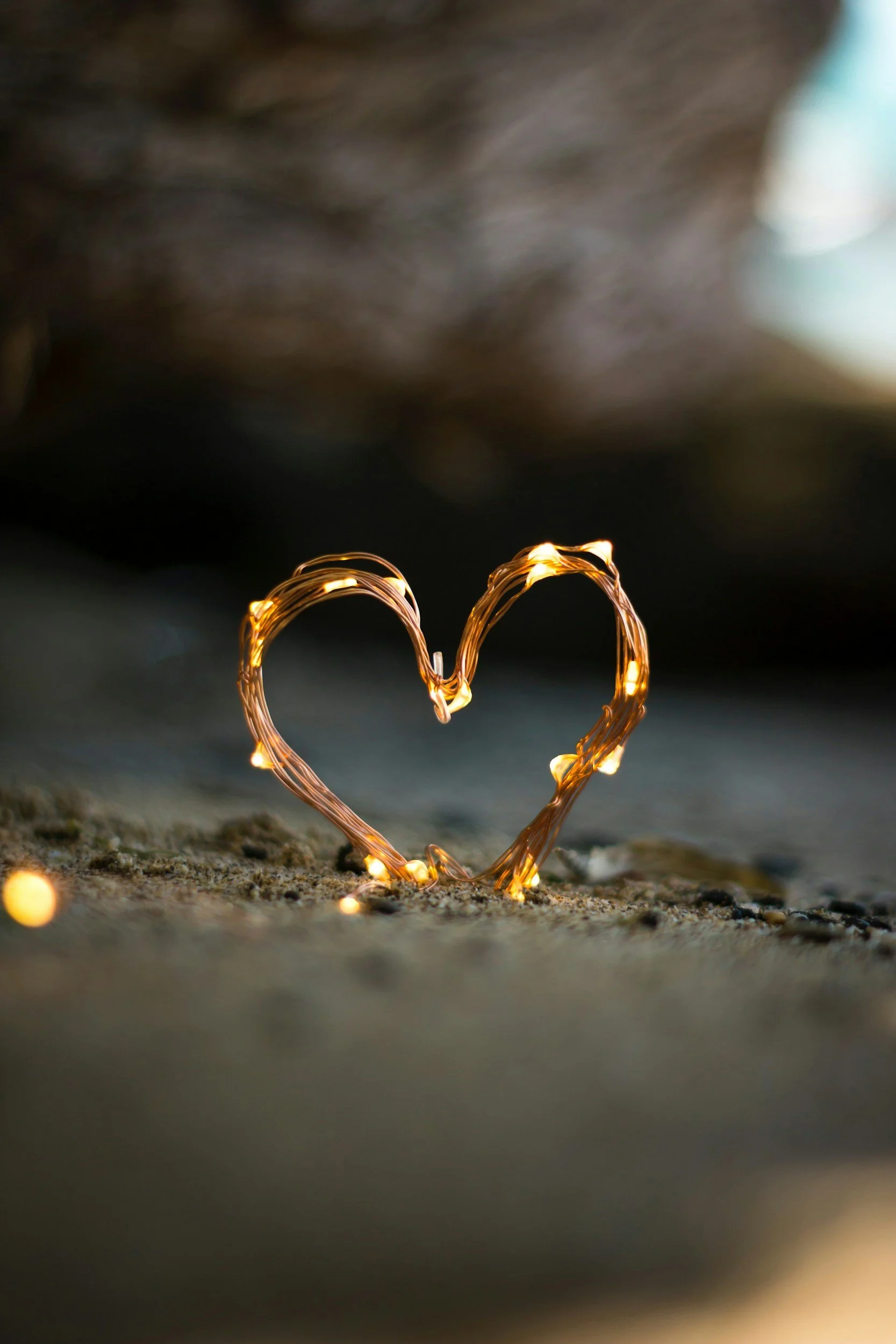 Close-up of a heart-shaped wire sculpture illuminated by small fairy lights, standing on a sandy surface with a blurred background.