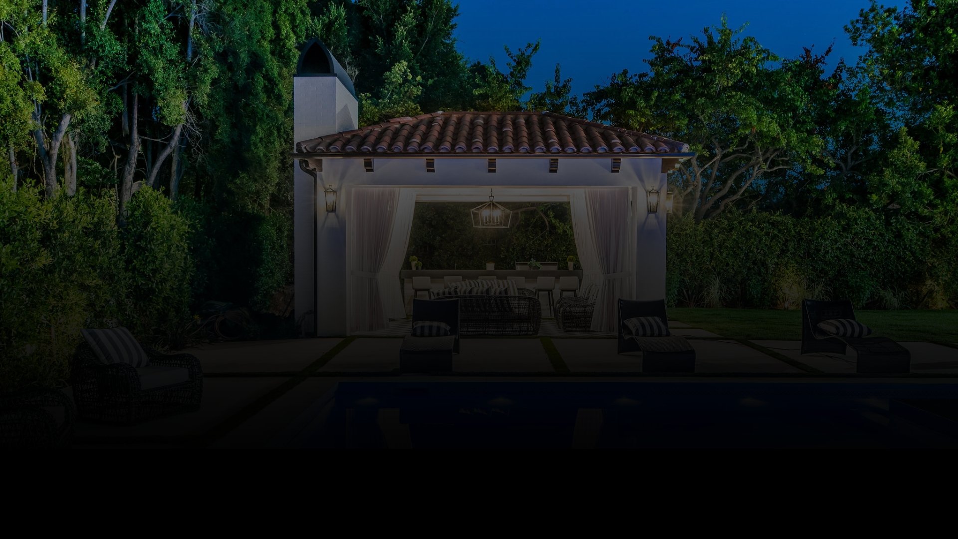 Nighttime backyard scene with a white outdoor pavilion, string lights, patio furniture, and lush trees surrounding the area.