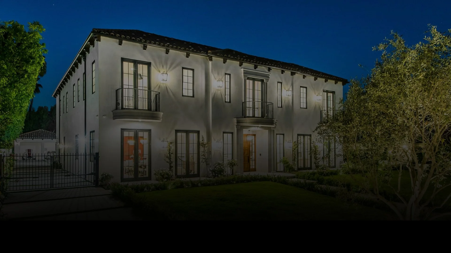 A two-story house illuminated at night with warm exterior lighting, featuring multiple tall windows, small balconies, and a front door, surrounded by greenery and trees.