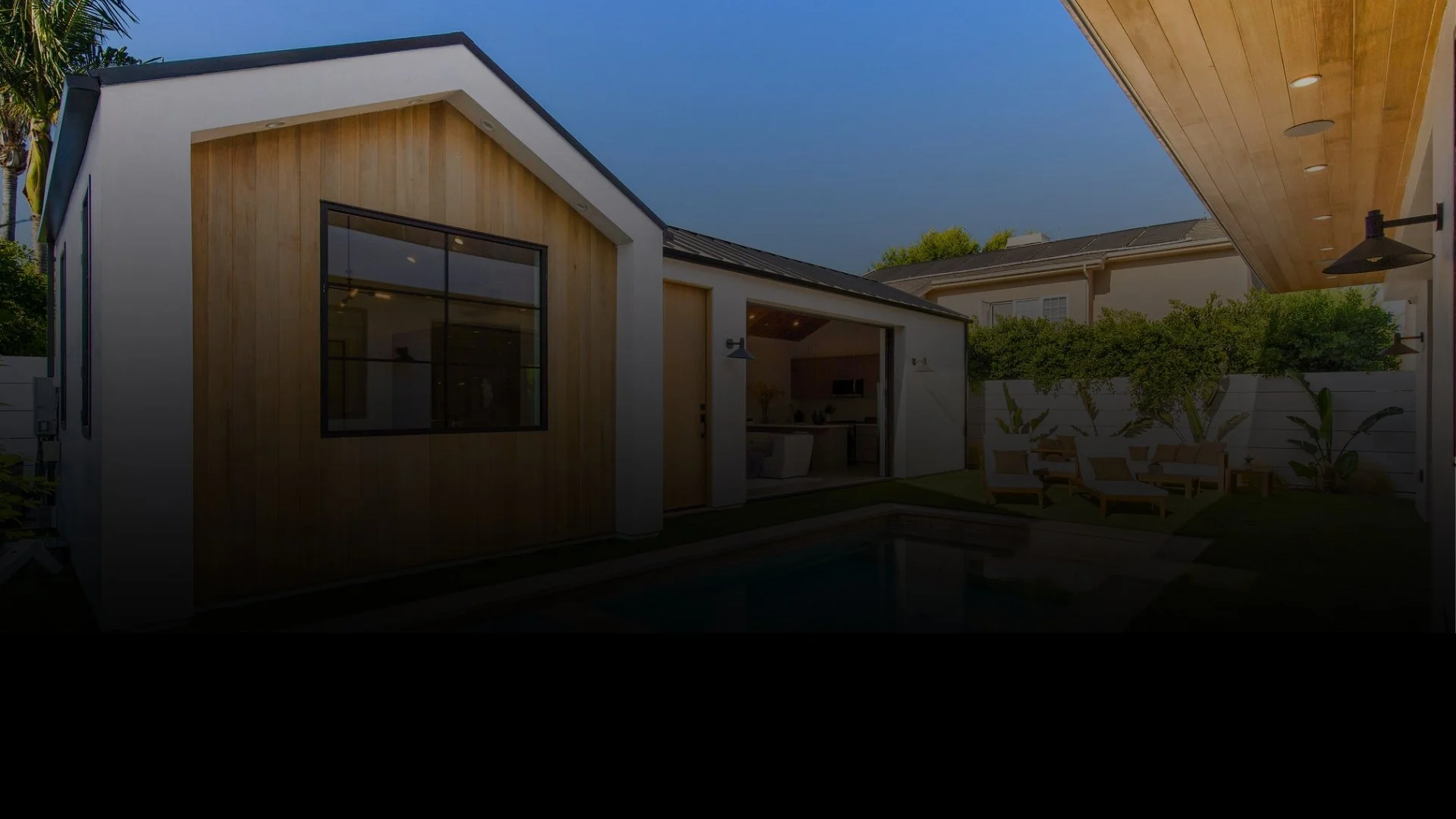 Modern residential backyard with a small pool, outdoor seating area, and a small structure with wood paneling and large windows, surrounded by a fence and greenery.