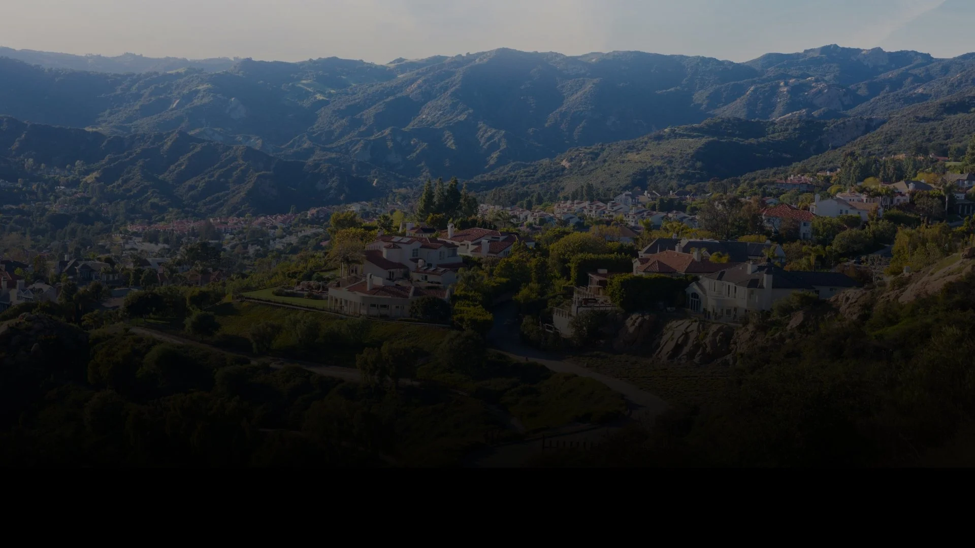 Houses nestled in a lush green hilly area, with mountains in the background under an overcast sky.
