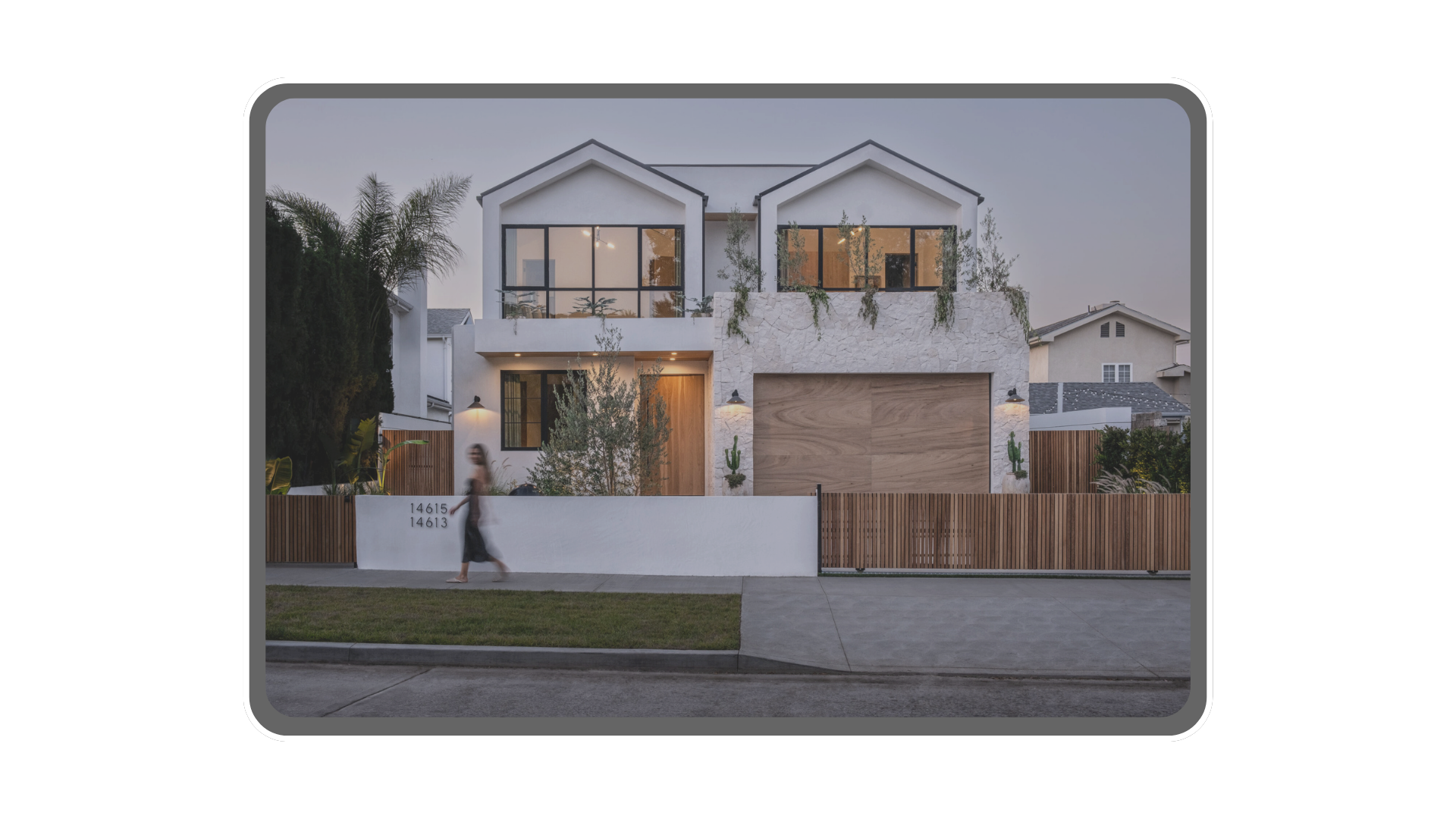 Modern white house with large glass windows, wooden garage door, and a concrete fence with house numbers 14613 and 14615, during dusk with a woman walking on the sidewalk.