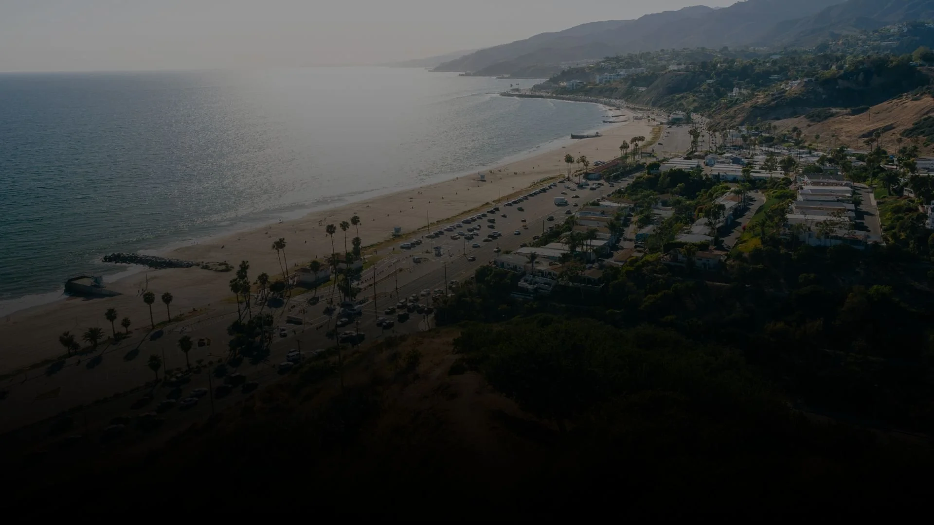 Aerial view of a beach with palm trees, parking lot, and residential houses along the shoreline, with hills in the background and the ocean on the left.