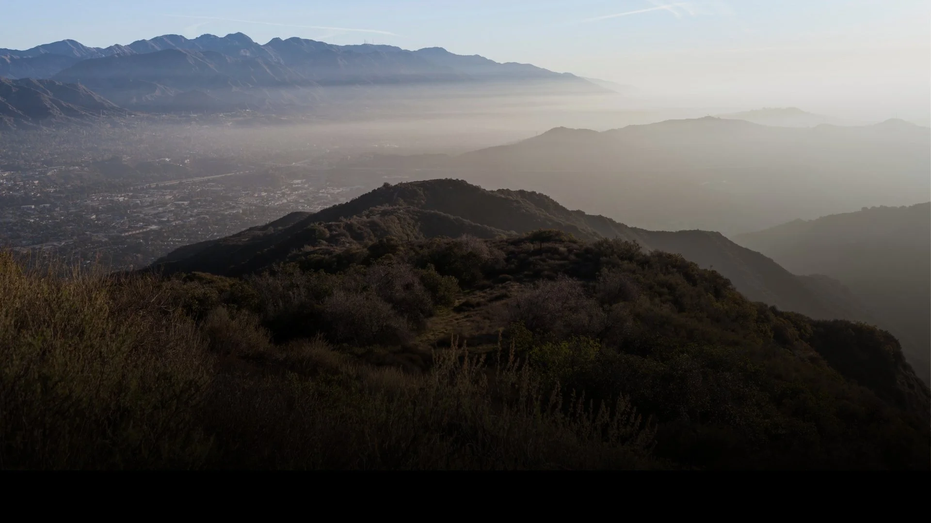 Mountain range with haze over a sprawling valley and urban area, overlooking rolling hills in the foreground.