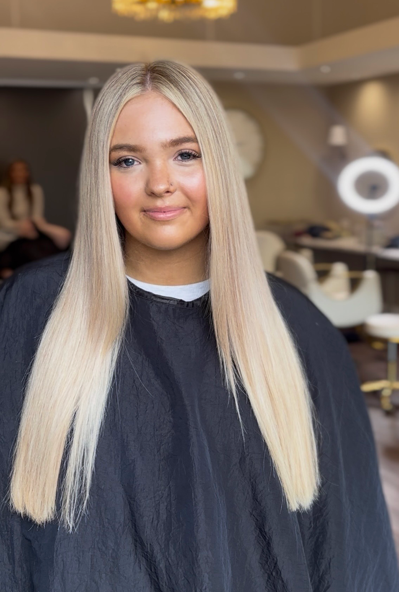 Young blonde woman in a salon with long straight hair, wearing black salon cape