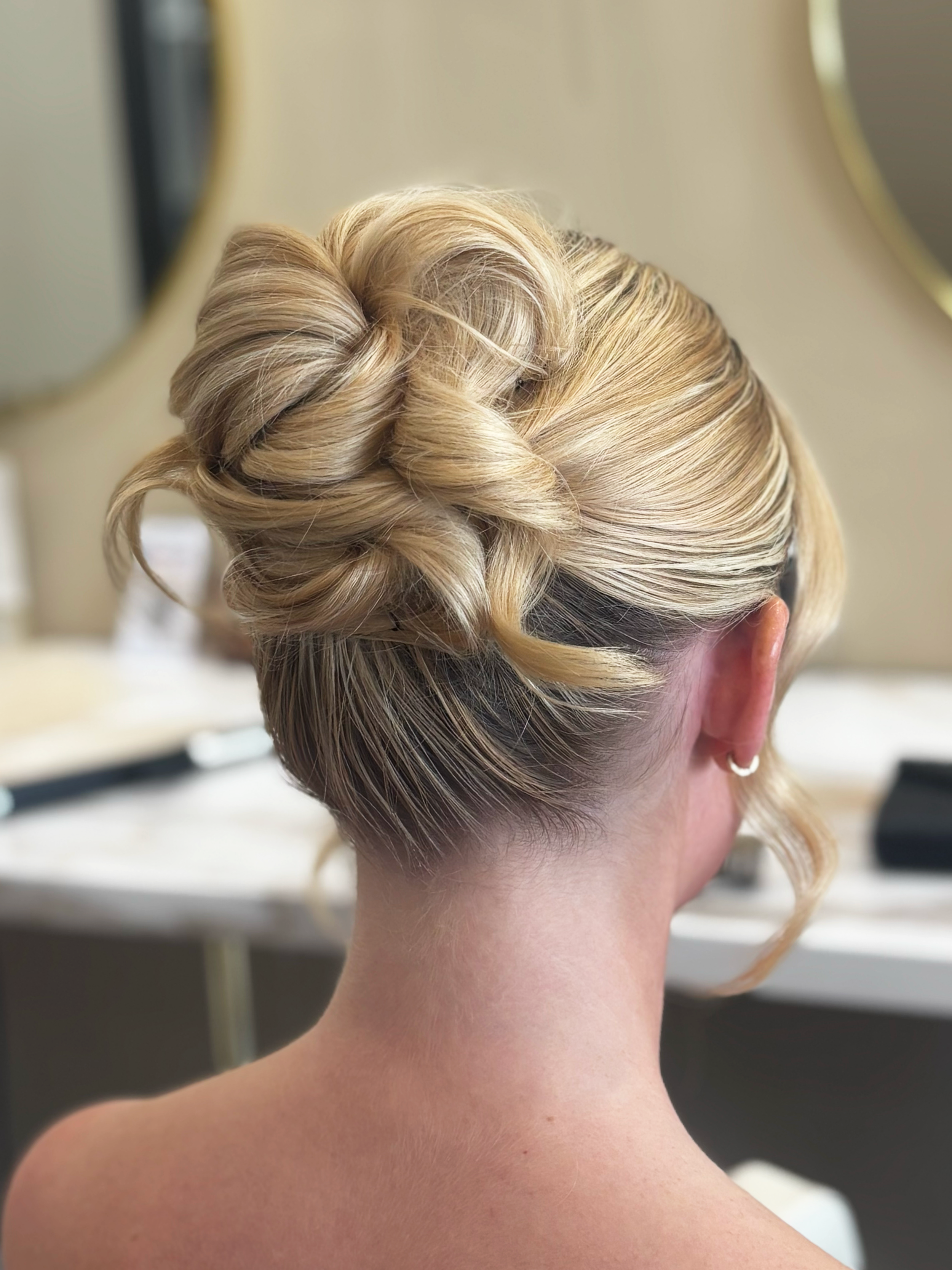 Back of a person's head with blonde, styled hair in an elegant updo, showing the neck and shoulder area
