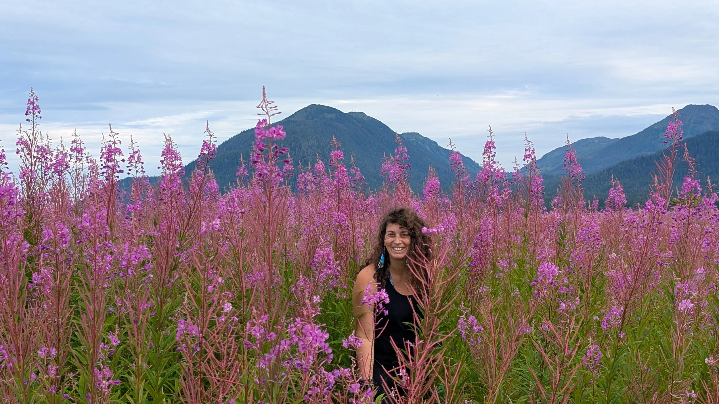 A woman with curly dark hair smiling in a field of tall pink flowers with mountains and a cloudy sky in the background.