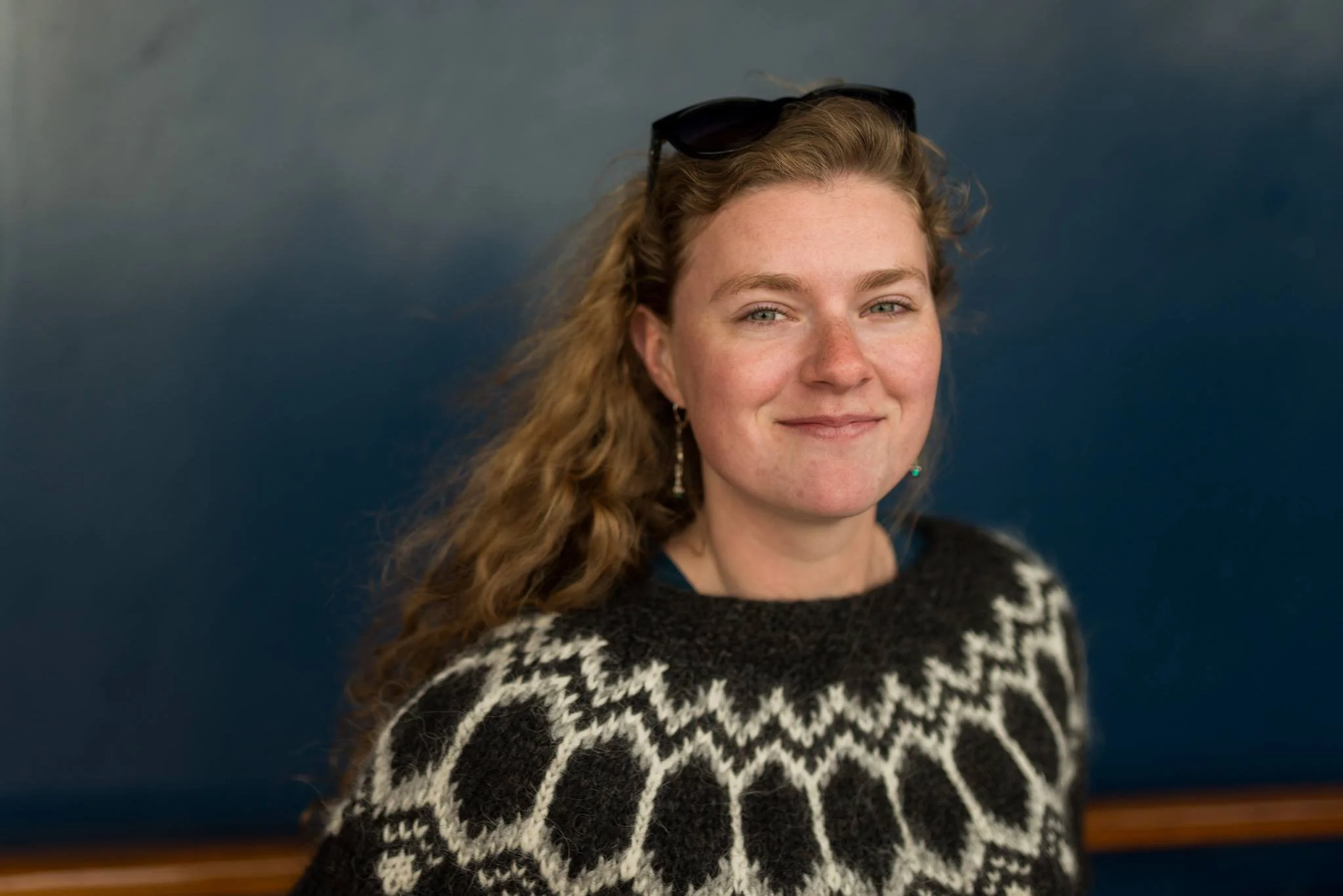 A young woman with long, curly red hair, lightly freckled skin, and blue eyes, smiling while wearing sunglasses on her head and a black and white patterned sweater, with a blurred dark blue background.