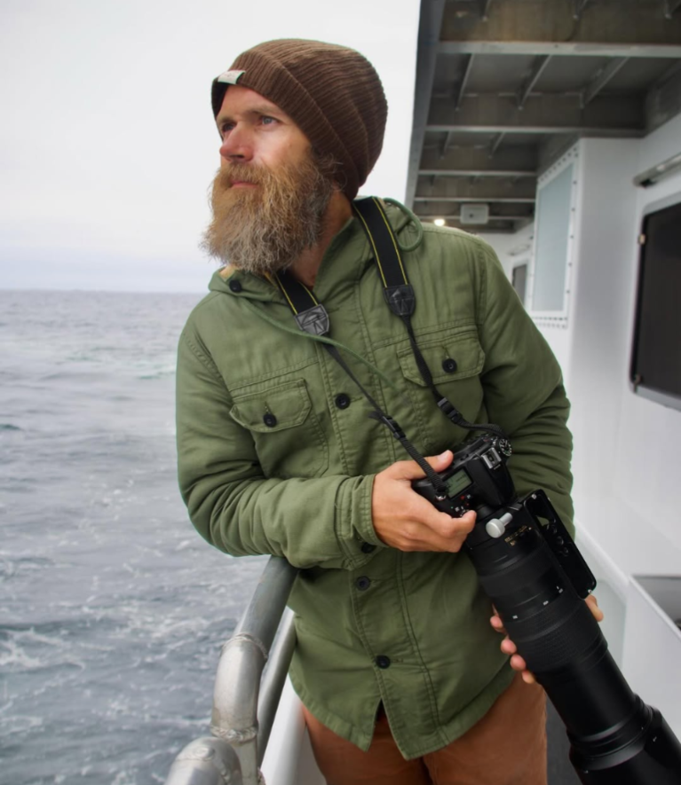 A man with a beard and brown knit cap standing on a boat, holding a camera with a large telephoto lens, looking out over the ocean.