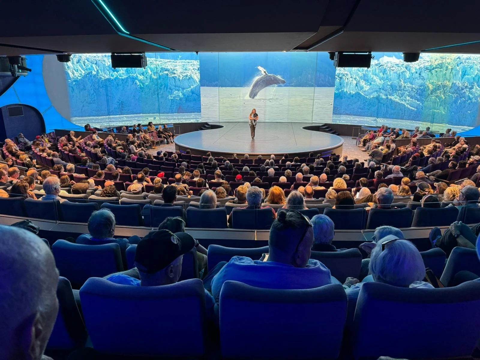 Audience watching a presentation at a large indoor auditorium with a stage projecting an image of a whale jumping out of the water.