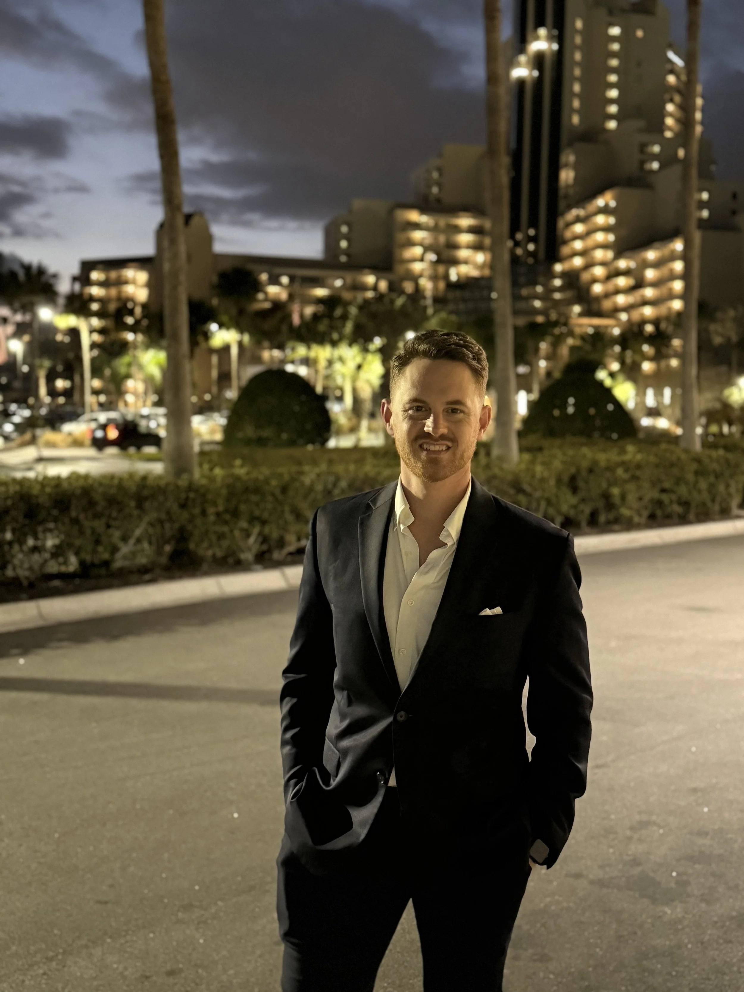 A man in a black suit and white shirt standing outdoors at night with tall buildings and palm trees in the background.