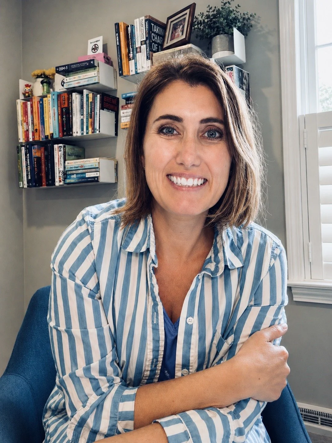 A woman with shoulder-length brown hair smiling at the camera, wearing a blue and white striped shirt, in a room with bookshelves and a window.