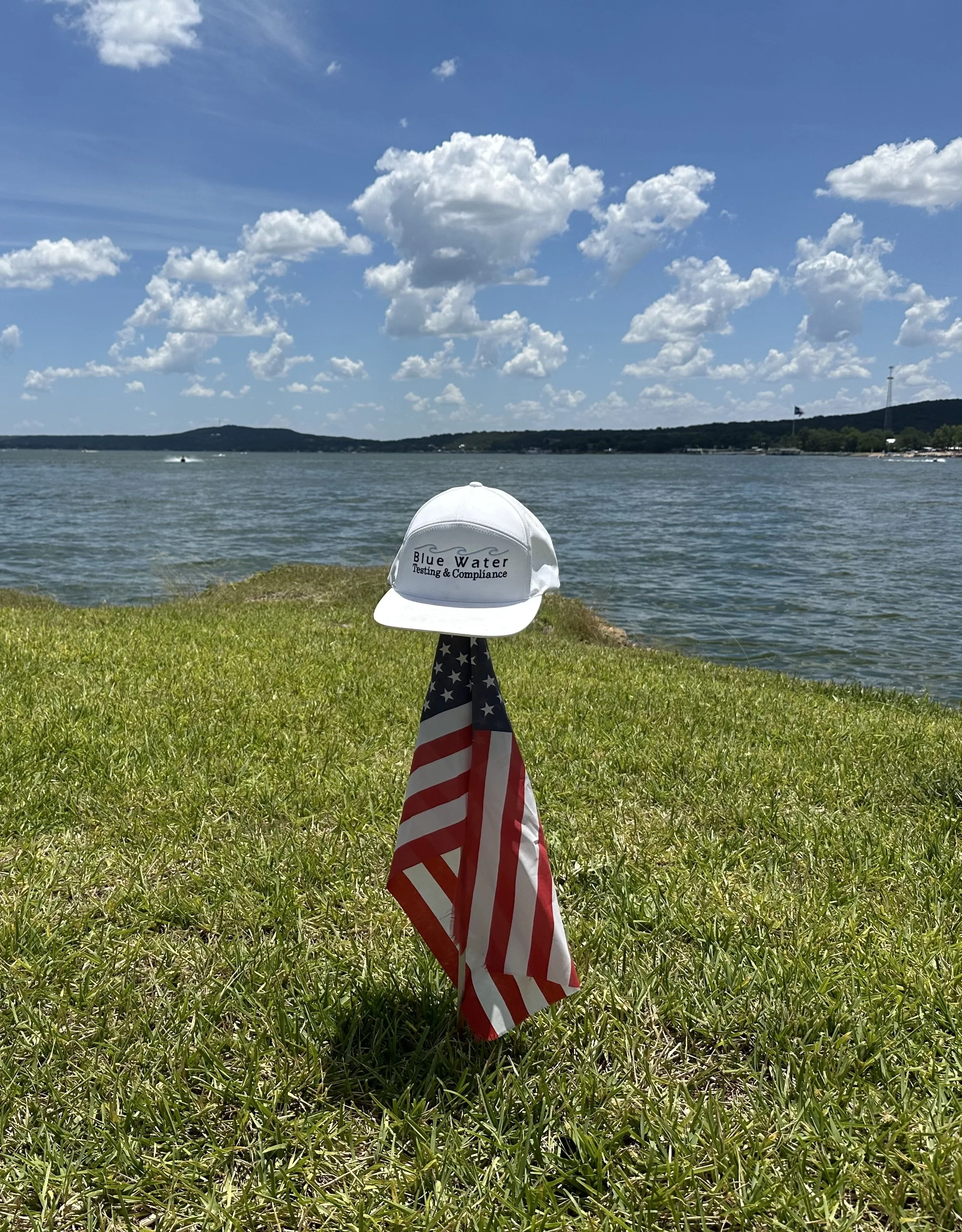 A white cap with the logo and text 'Blue Water Testing & Compliance' on a grassy area near a body of water. The background features a blue sky with scattered clouds and distant hills.