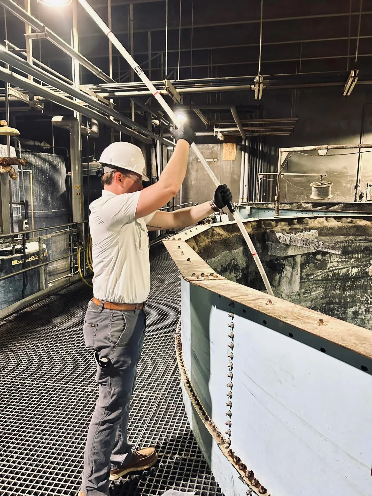 A worker in safety gear, including a hard hat and gloves, is inspecting or working on industrial equipment, possibly inside a manufacturing or processing facility with metal pipes and structural framework.