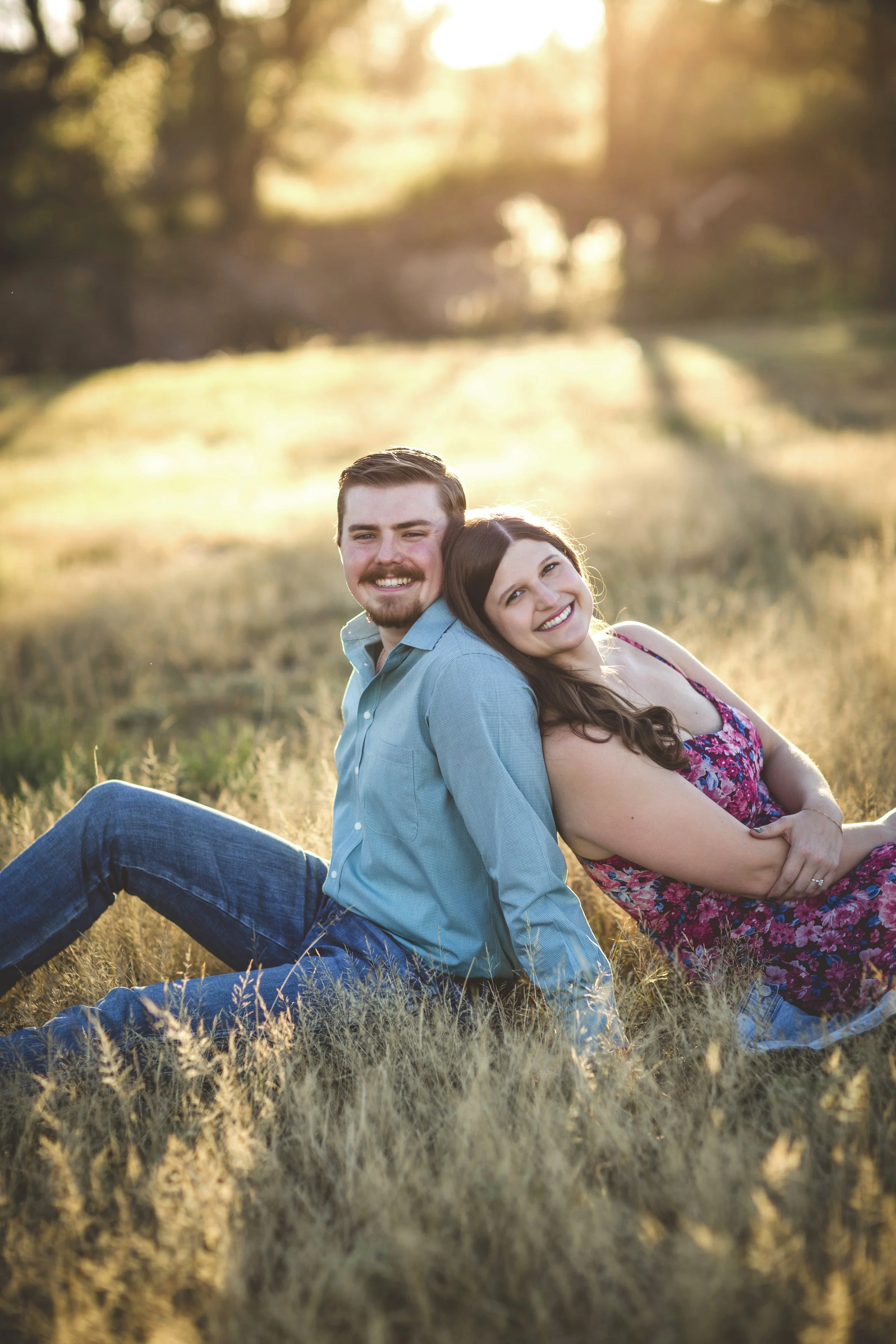 A happy couple sitting on the grass in a field during sunset, with the man wearing a light blue shirt and the woman in a floral dress, smiling and leaning back against each other.