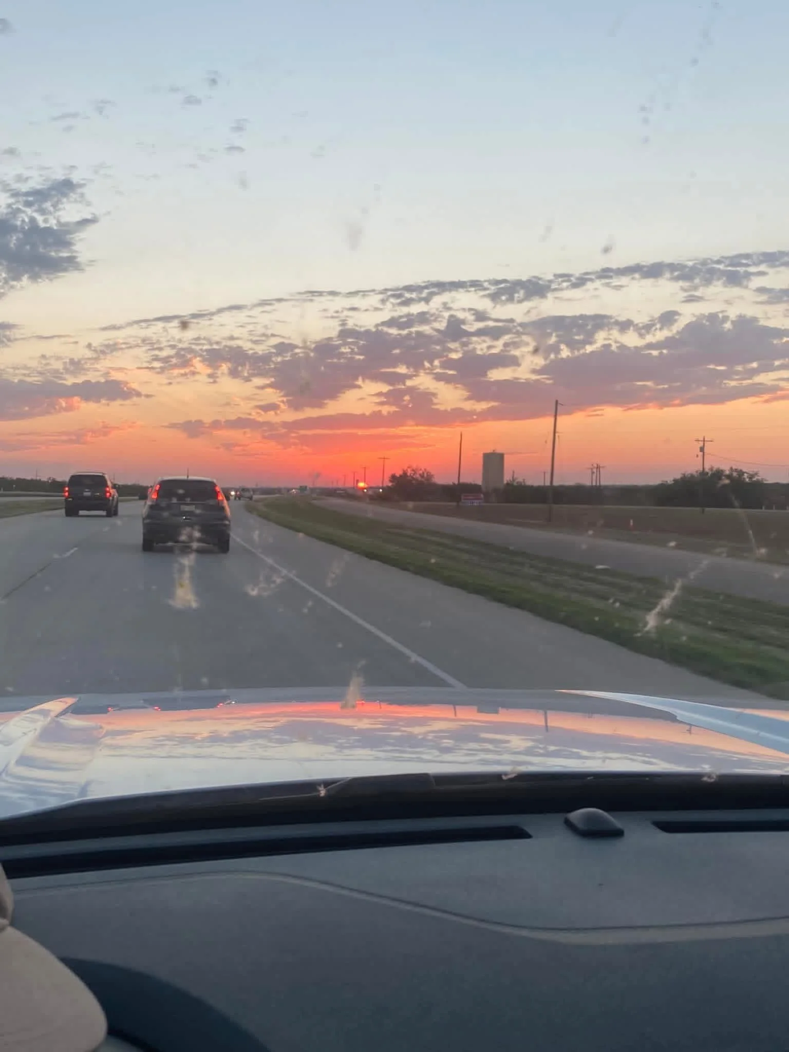 View of a sunset sky with scattered clouds and a highway with several cars driving toward the horizon, seen from inside a vehicle.