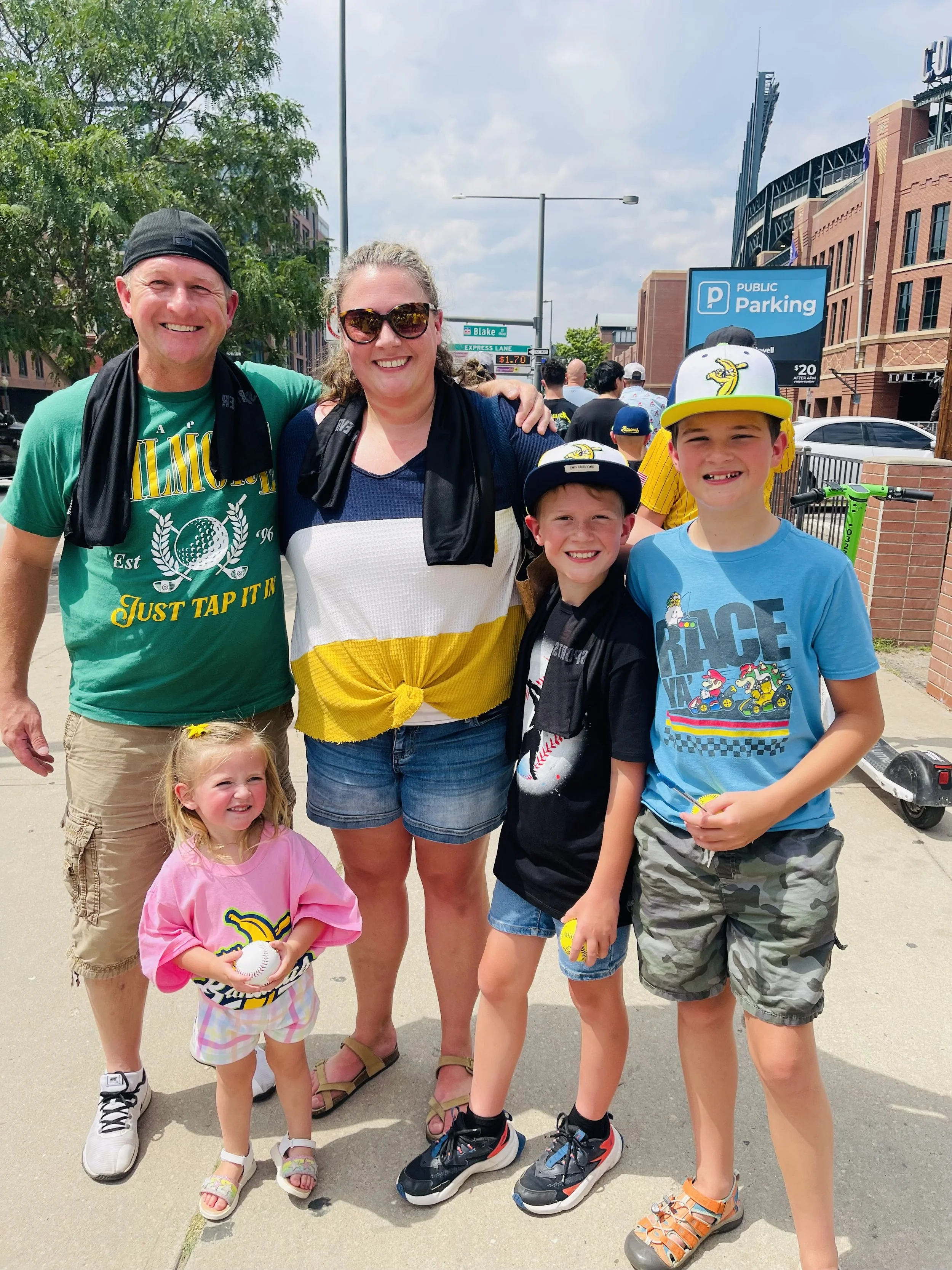 A group of people standing outside near a parking lot, including two adults and three children, with a baseball logo on the children's clothing, and a sign indicating public parking in the background.