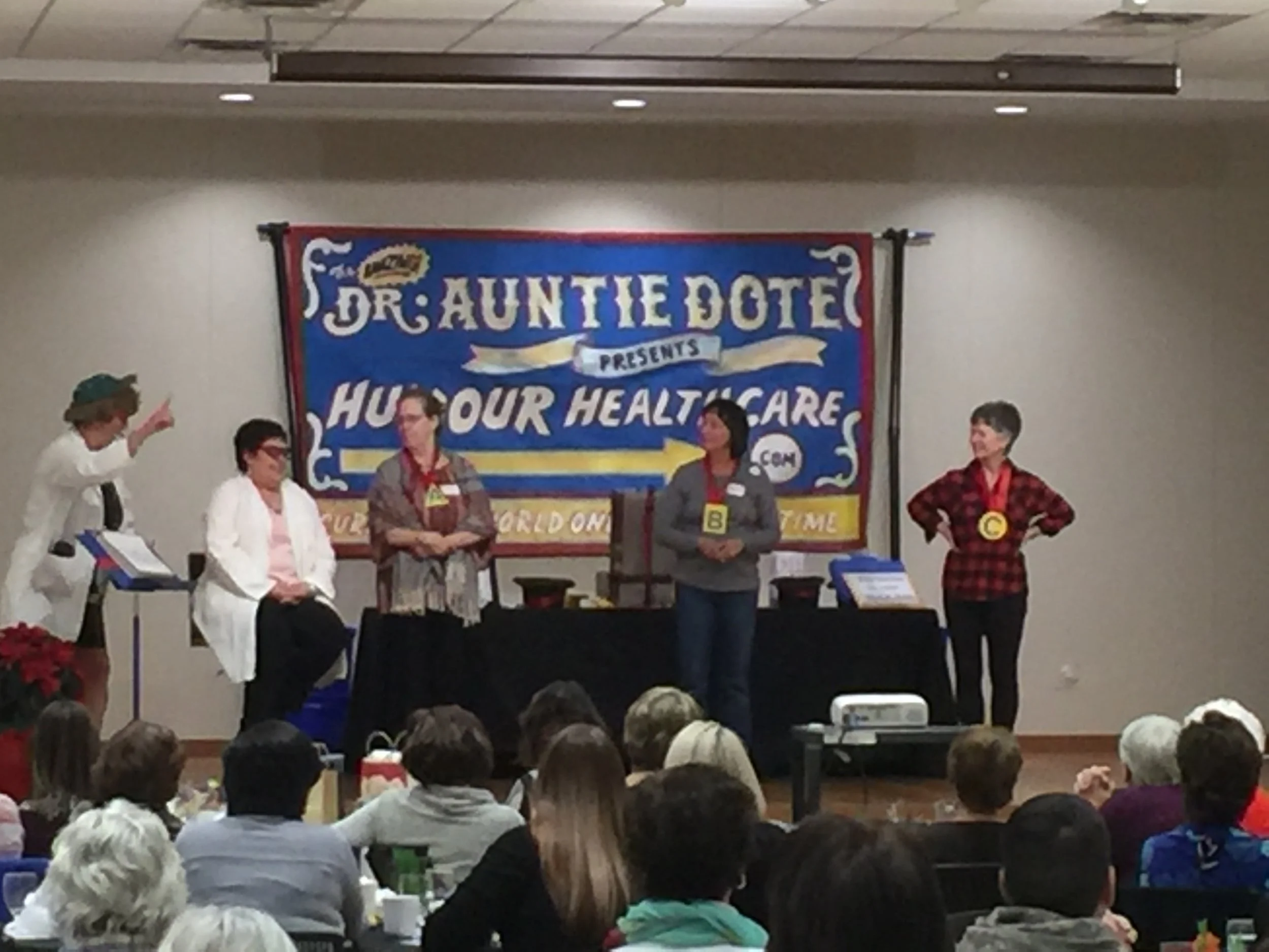 Heather D. Swain as Dr. Auntie Dote she is giving a keynote speech. Humour Healthcare sign is on the wall behind the volunteers