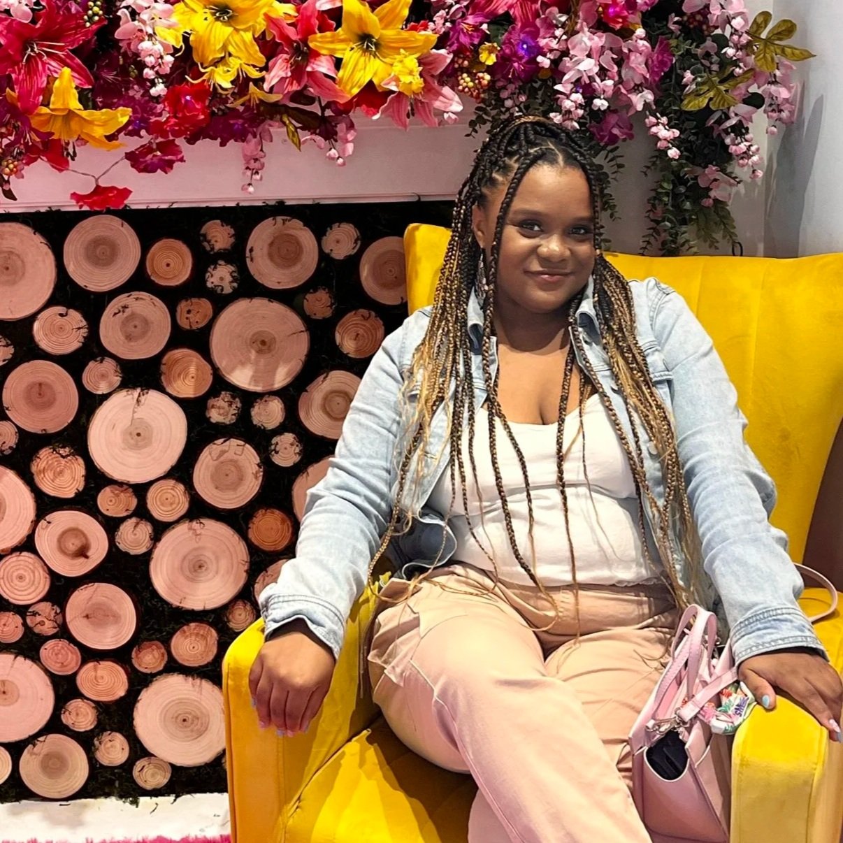 A woman with braided hair sitting on a yellow armchair, smiling, with a background of colorful flowers and stacked wood logs.
