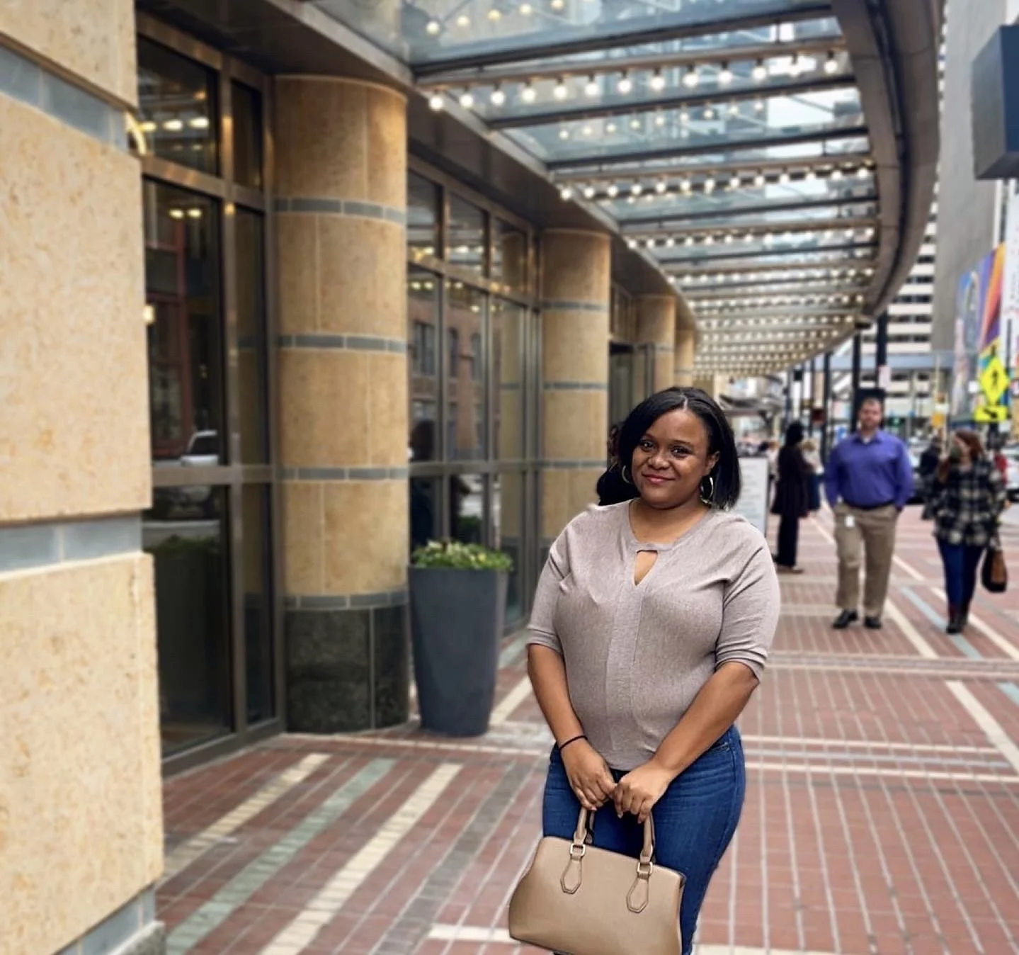 A woman with black hair standing on a brick sidewalk outside a building with large glass windows and a metal awning, holding a beige handbag.