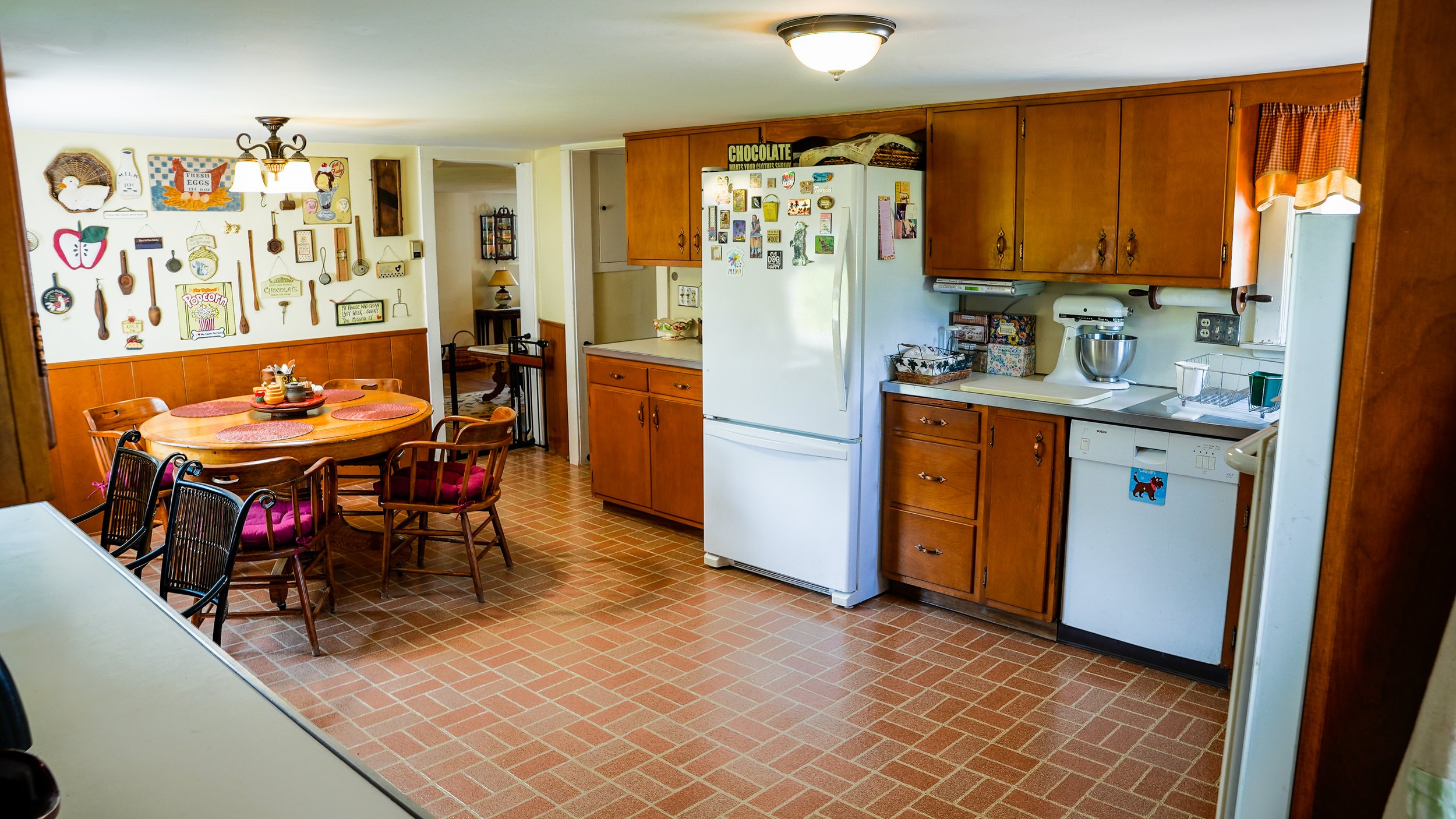 A cozy kitchen with wooden cabinets, white appliances, and a tiled floor. The dining area features a wooden table with four chairs, and the wall is decorated with various kitchen-themed decorations.