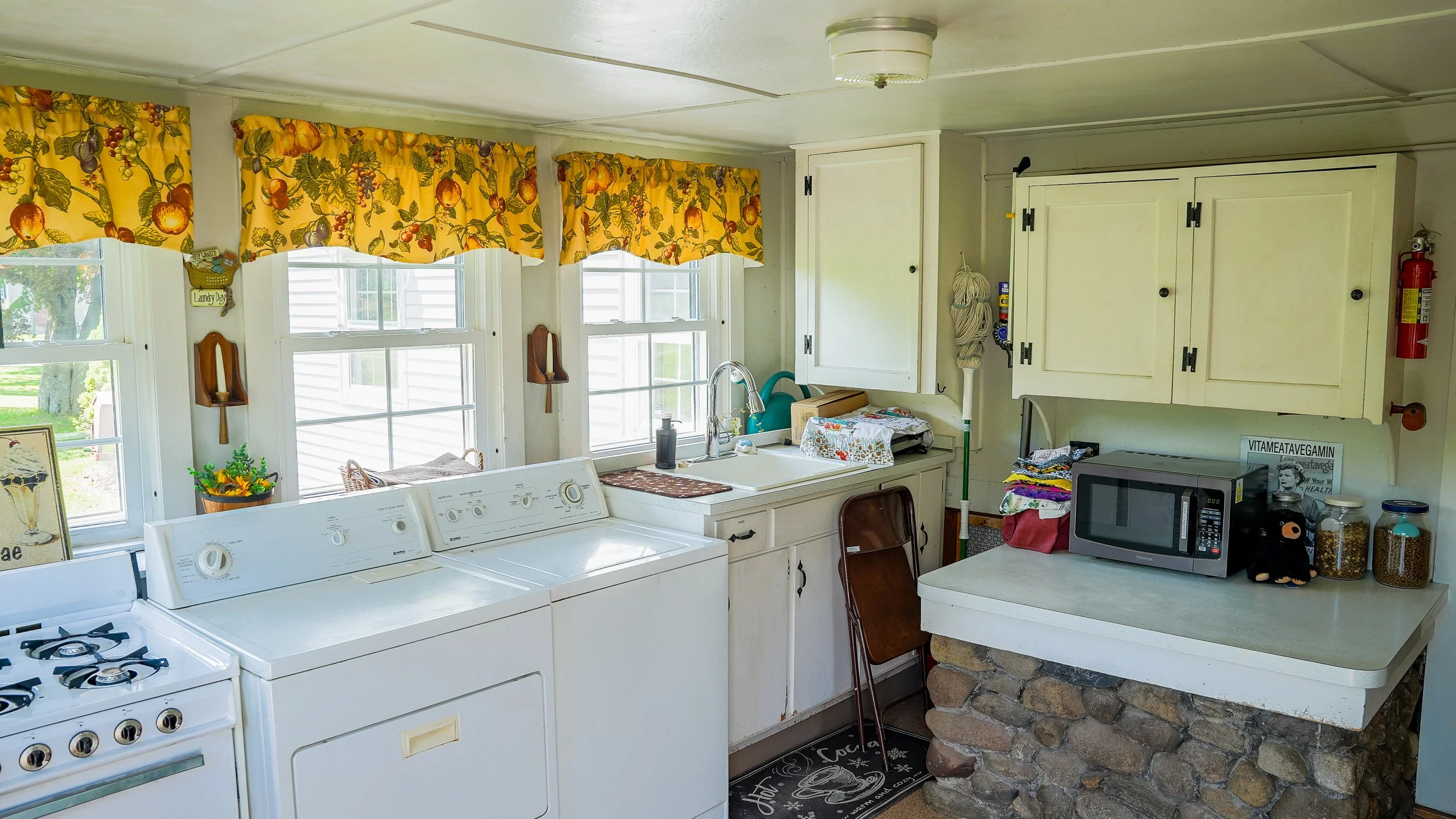 A kitchen with multiple white windows covered by yellow valances with fruit and leaf pattern. There are two white washing machines in front of the windows, and a white sink with a green watering can and paper towels nearby. To the right, there are wooden cabinets, a microwave, jars of grains or spices, and a small table with a patterned mat. A brick and stone structure supports a countertop, with a chair in front and a black mat on the floor.