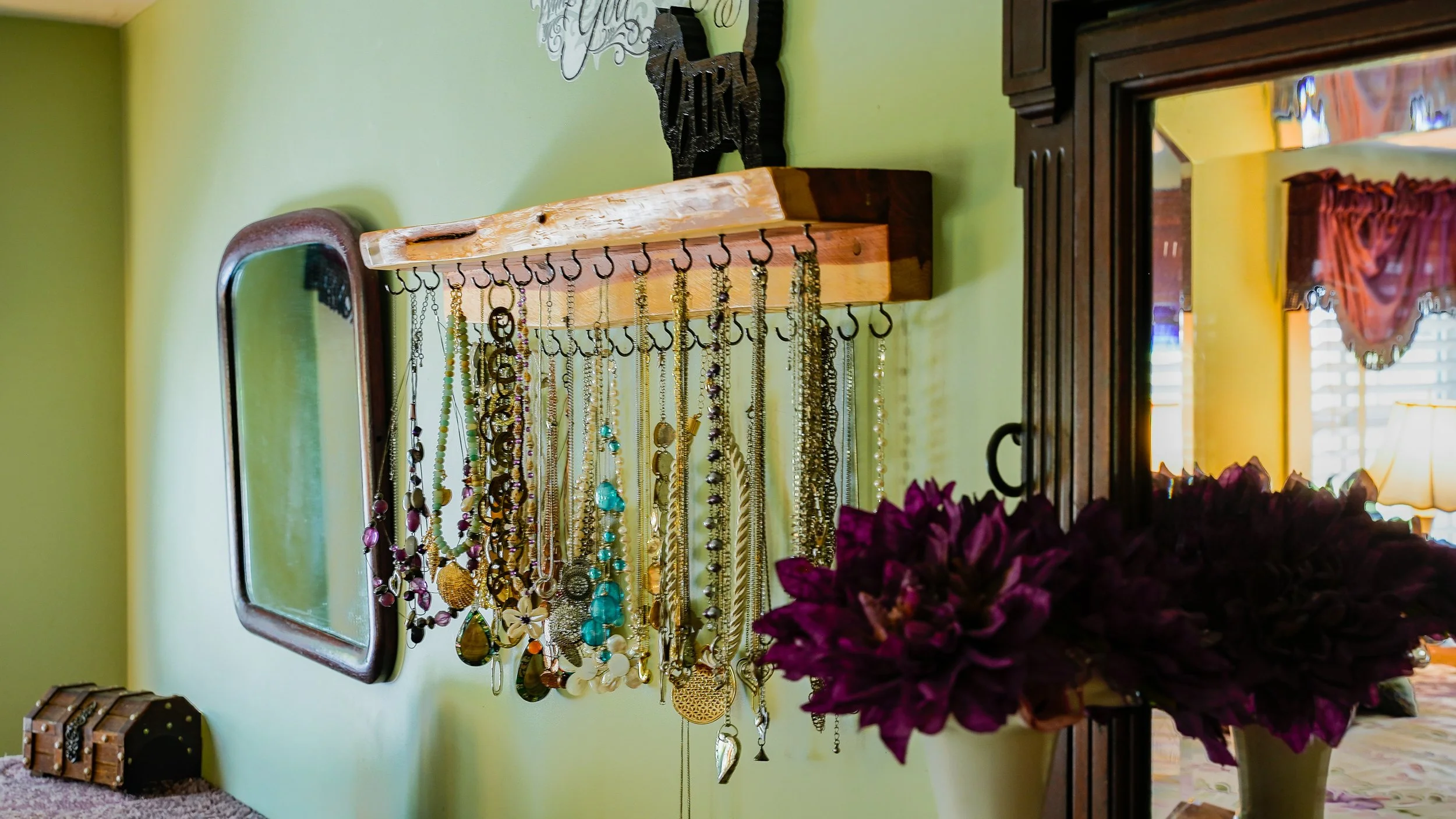 Jewelry display with various necklaces hanging on hooks against a yellow wall, with a mirror and a vase of purple flowers nearby.