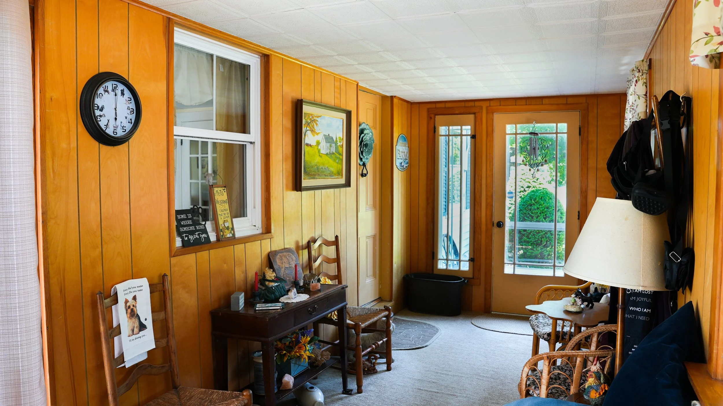 Living room with wood-paneled walls, a clock, framed pictures, chairs, a door with a screen door, and large windows with curtains showing greenery outside.