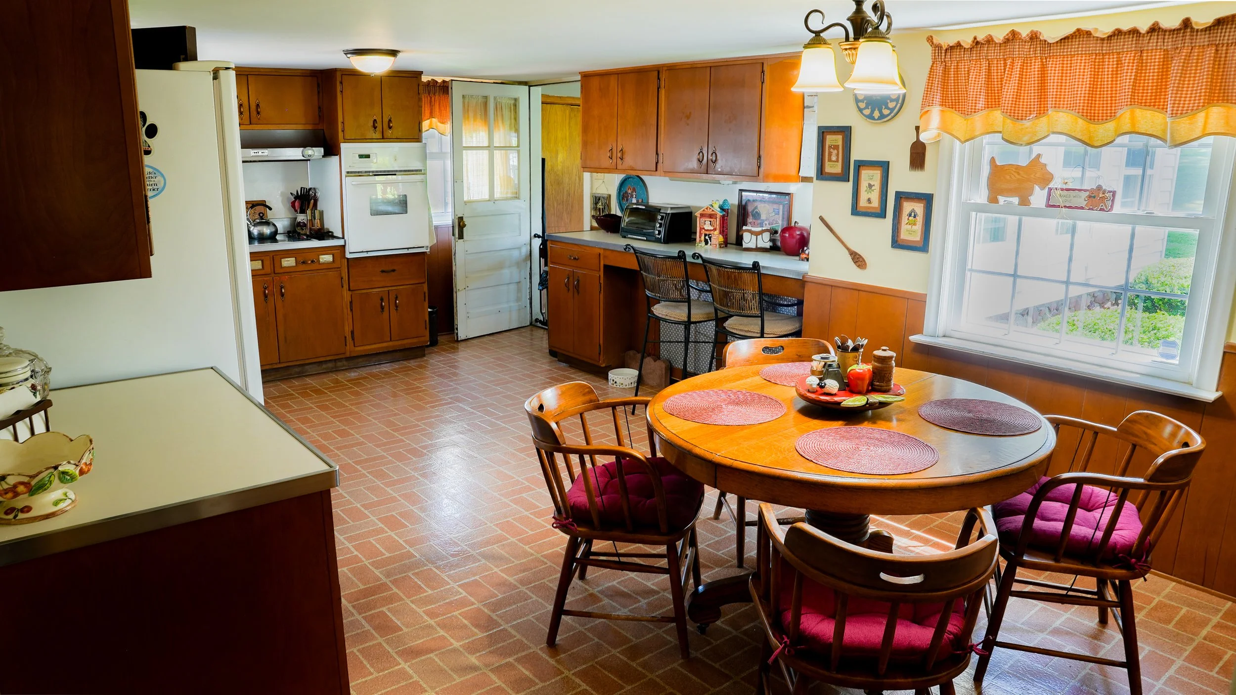 Kitchen with wooden cabinets, a round dining table with red placemats and chairs, and a window with orange gingham curtains.