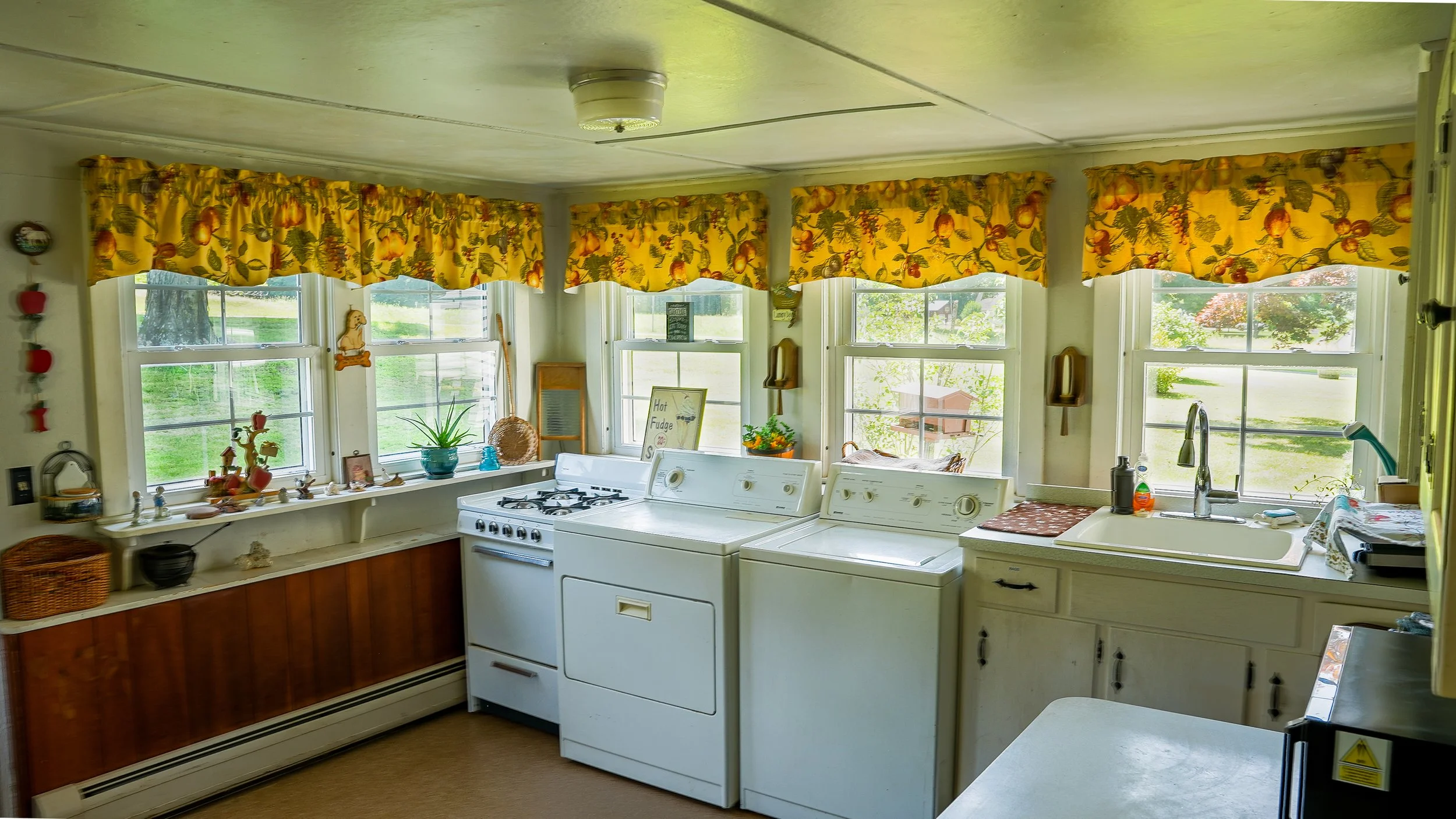 A kitchen with a row of windows with floral yellow curtains, a white stove, a white washing machine, and a sink with a faucet. Decor items and plants are on the window sill.