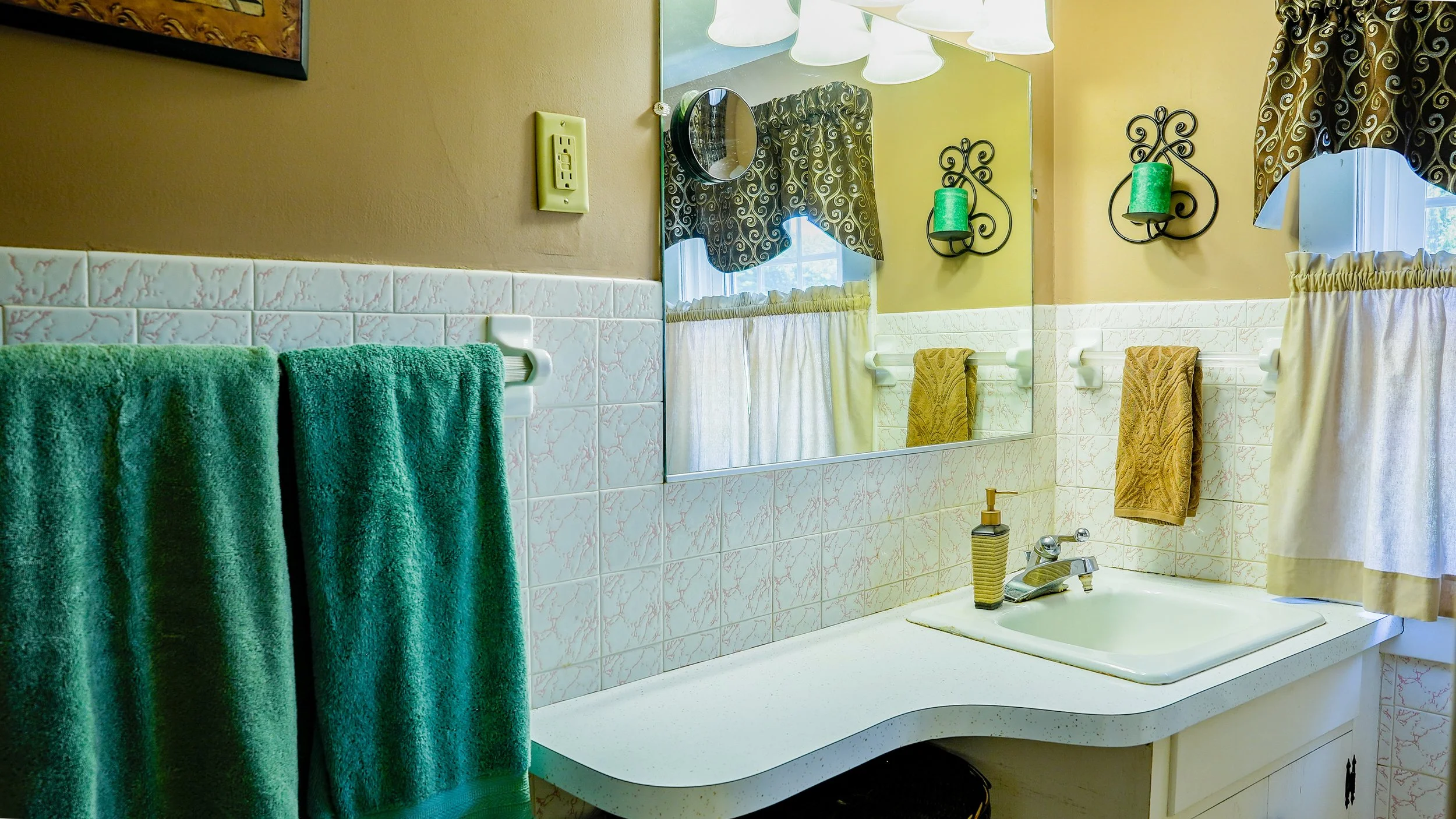 A bathroom with a mirror, green towels hanging on a towel rack, a window with curtains, a sink with a soap dispenser, and wall sconces with green candles.