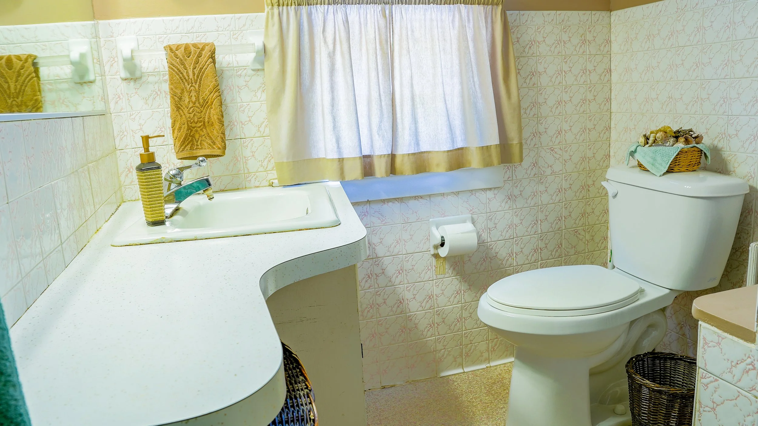 A vintage bathroom with a white sink, gold soap dispenser, mustard towel, beige curtains, toilet with a basket of seashells on top, and wicker laundry baskets.