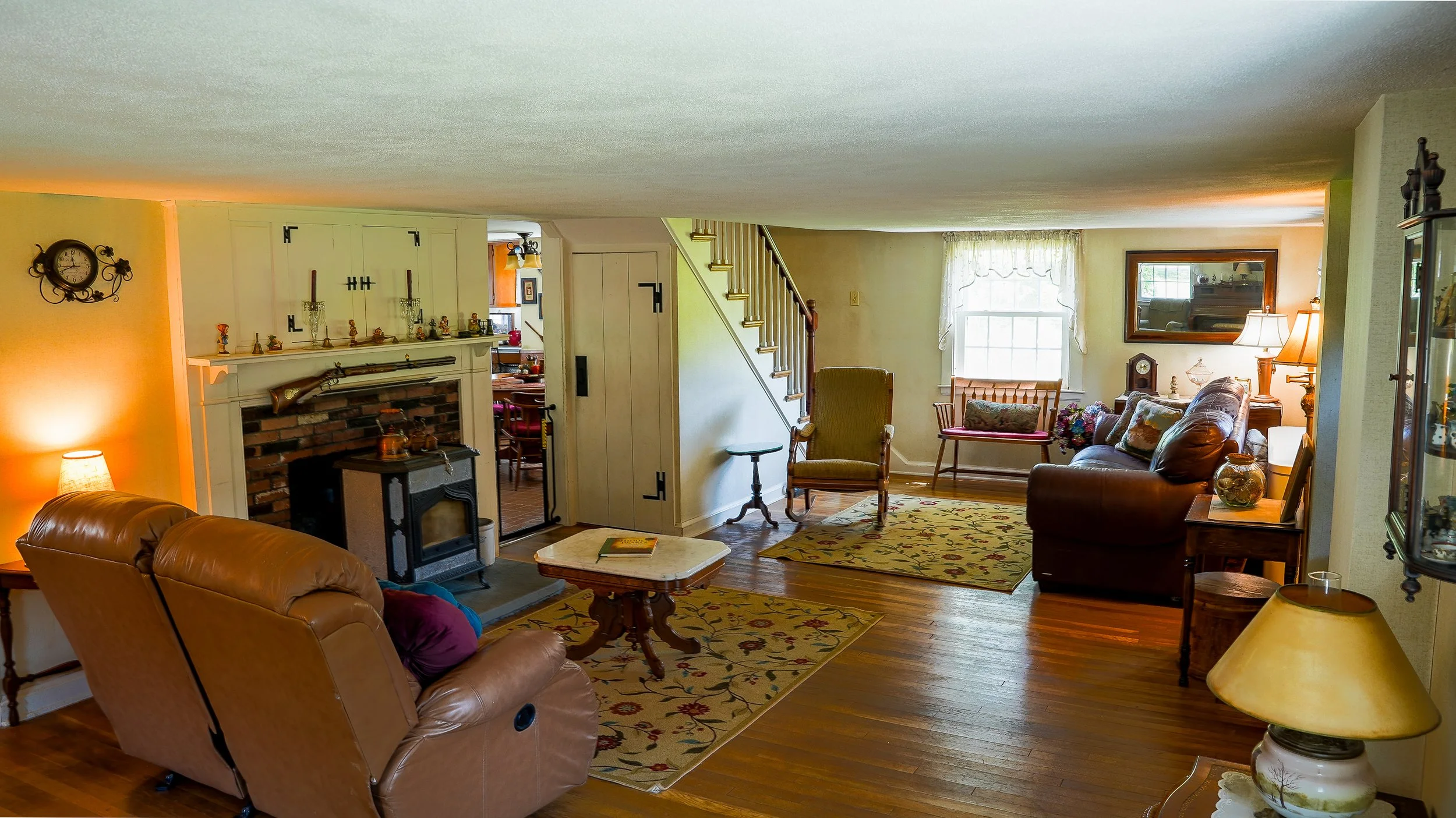 A cozy living room with tan leather sofas, a fireplace, a wooden coffee table, and a patterned area rug. There are windows with white curtains, a mirror on the wall, and various decorative items including lamps, a clock, and figurines.