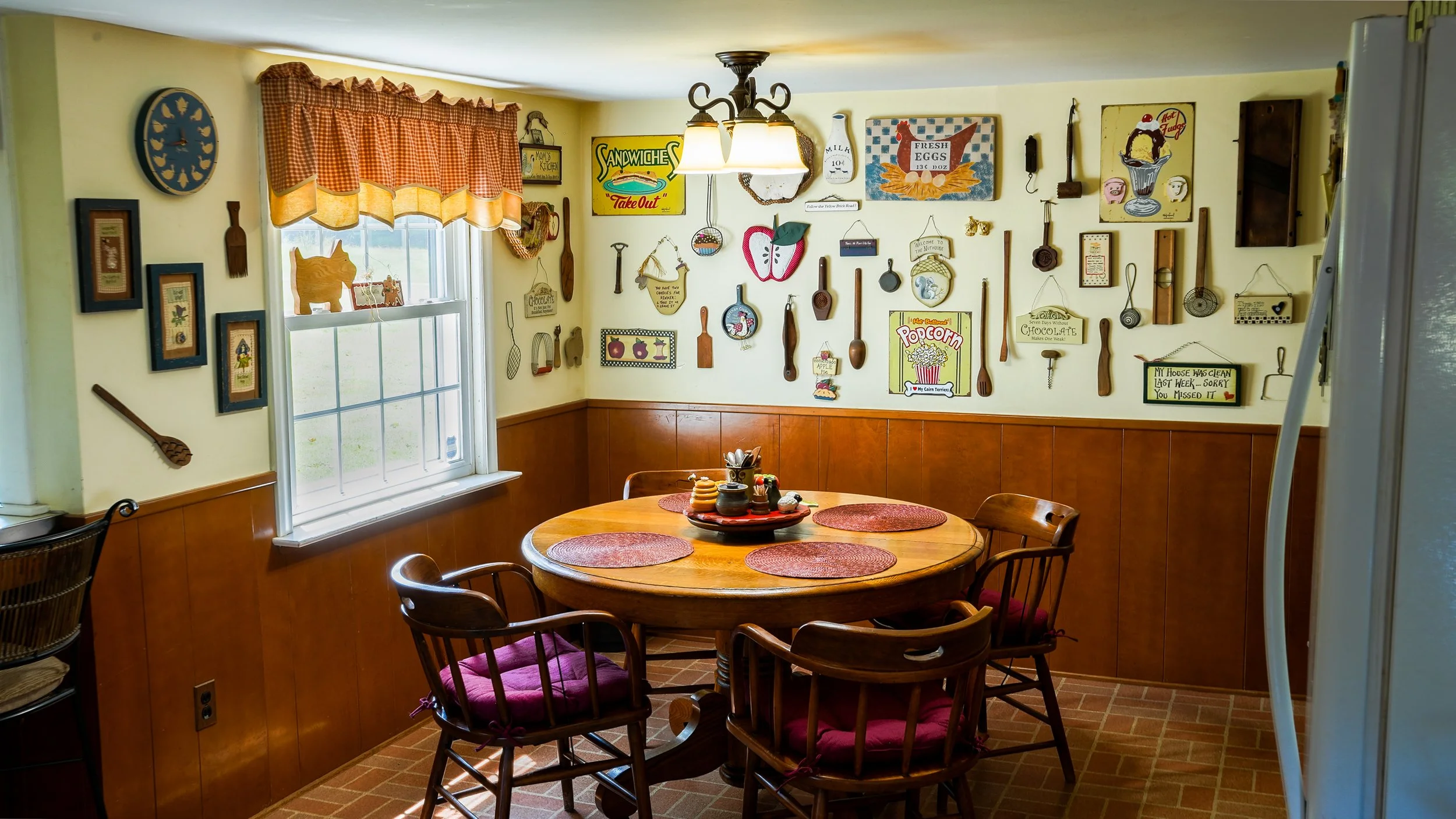 A cozy kitchen dining area with a round wooden table surrounded by six chairs with red cushions. The wall is decorated with vintage kitchen-themed signs, utensils, and artwork. There is a window with a red and white checkered curtain, and a ceiling light fixture above the table.