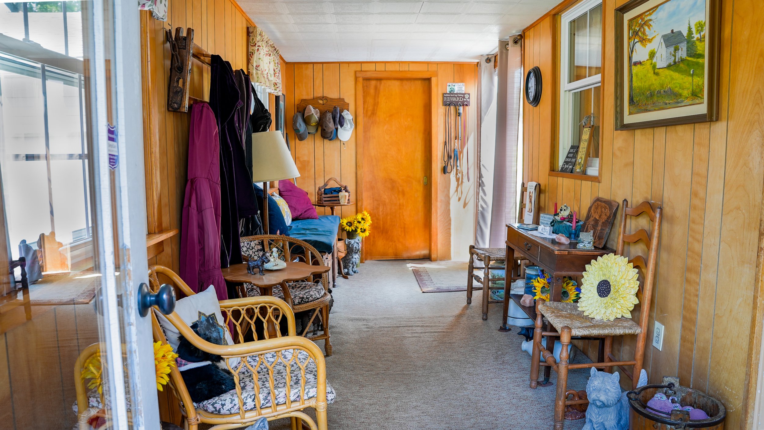A cozy living room with wood-paneled walls, decorated with hats, hats, and framed artwork, featuring a small wooden table, sunflower-based decorations, and various knick-knacks, illuminated by natural light from window blinds.