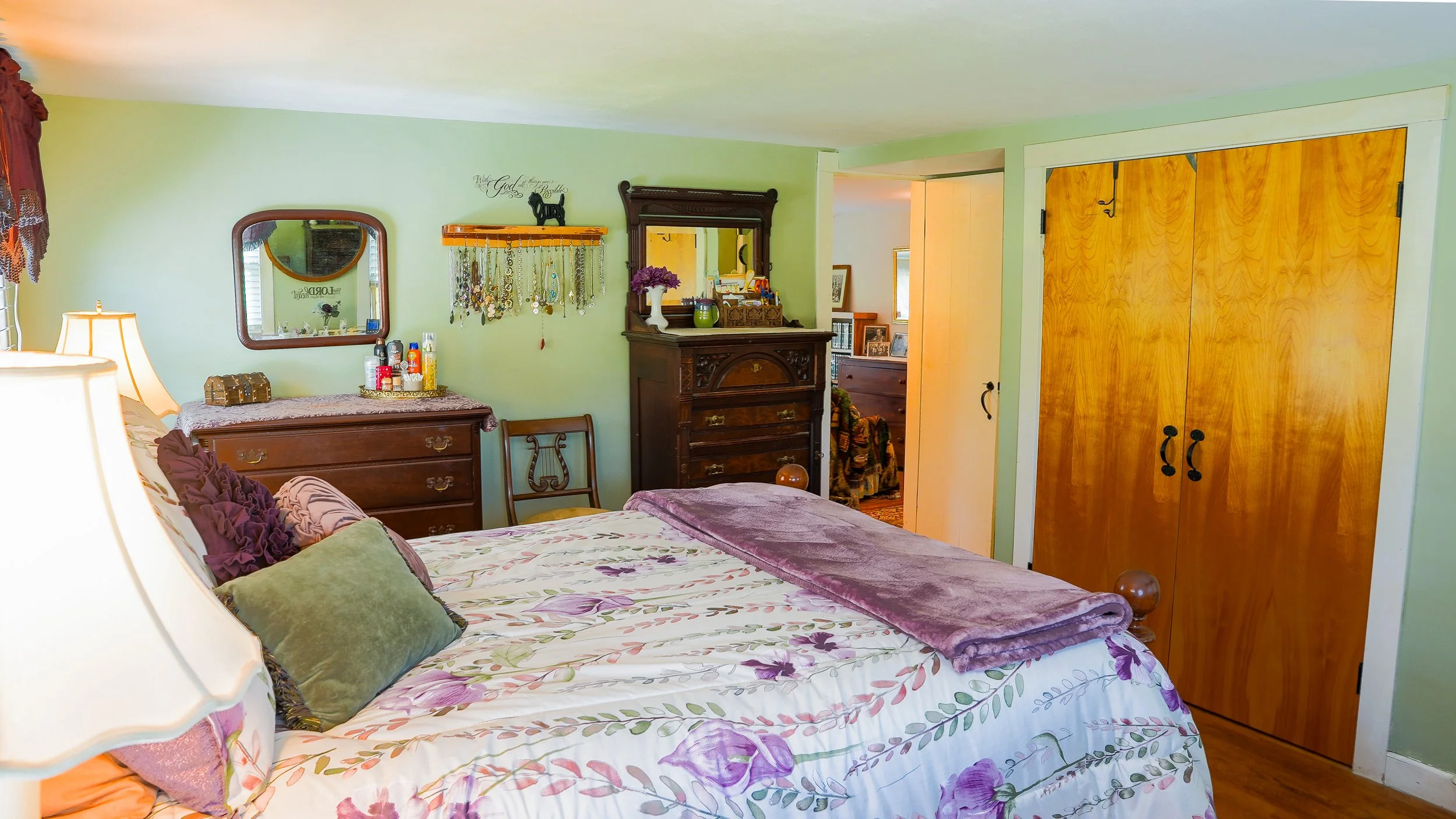 A bedroom with green walls and wooden furniture, including a bed with floral bedding and decorative pillows, a dresser with a mirror, and a closet with wooden doors. Various personal items and jewelry hang on the wall, and there is a small view into another room.
