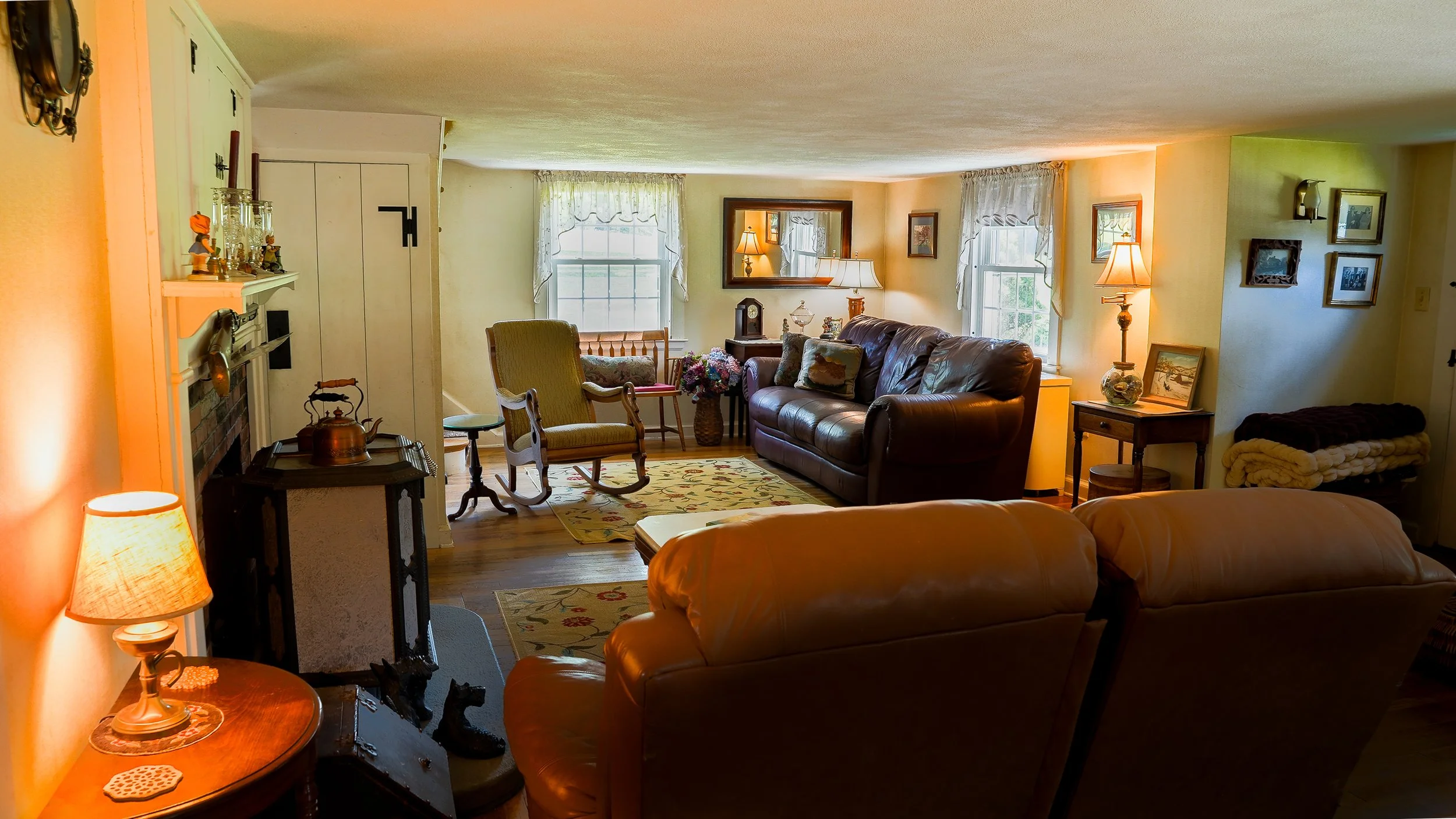 A cozy living room with natural light from multiple windows, featuring a leather sofa, vintage rocking chair, side tables with lamps, and framed artwork on the walls.