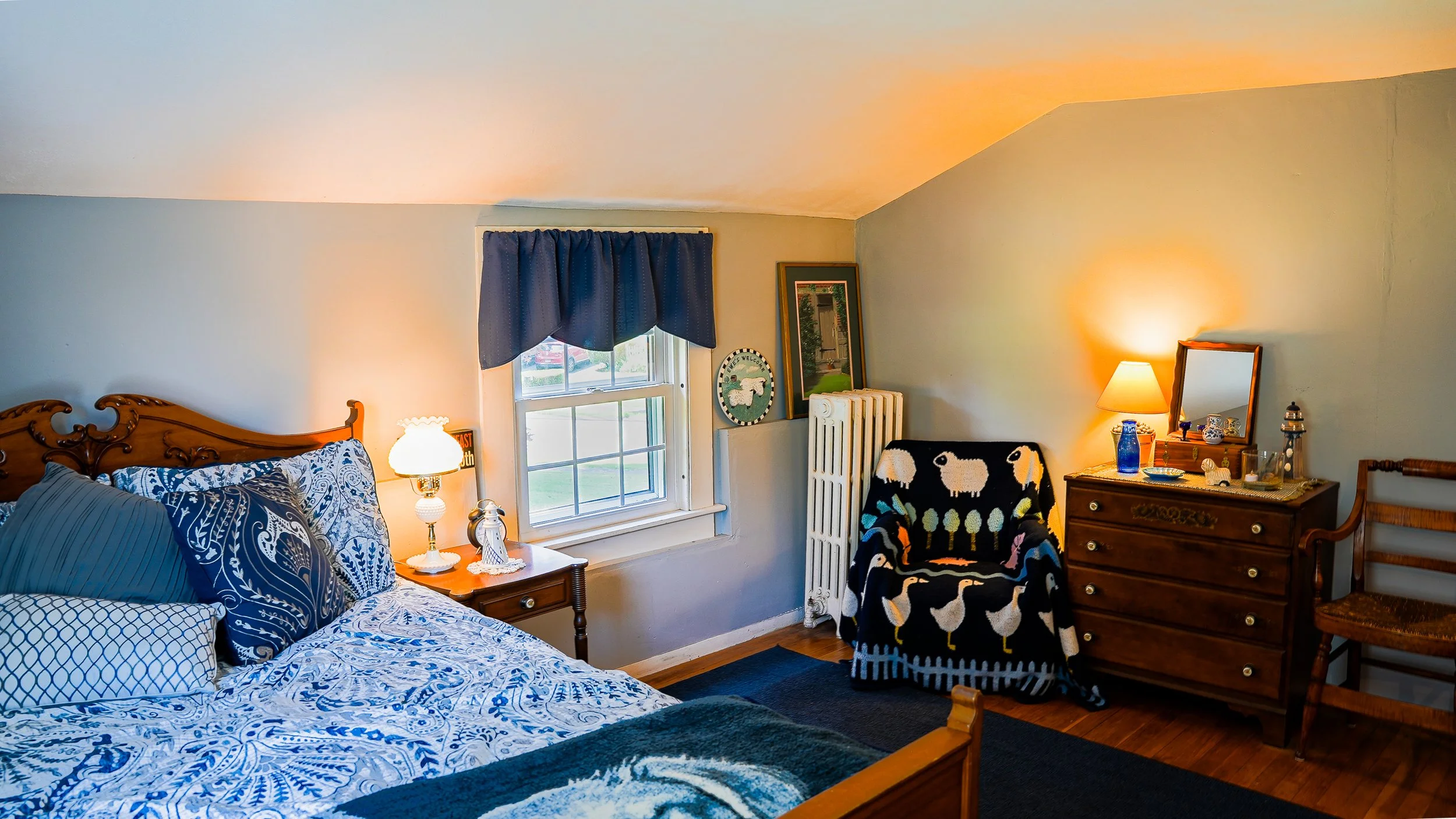 A cozy bedroom with a wooden bed, blue patterned bedspread, and pillows. A nightstand with a lamp and decorative items. A window with blue curtains. A chair with a black sheep blanket. A wooden dresser with a mirror, lamp, and decorative bottles.