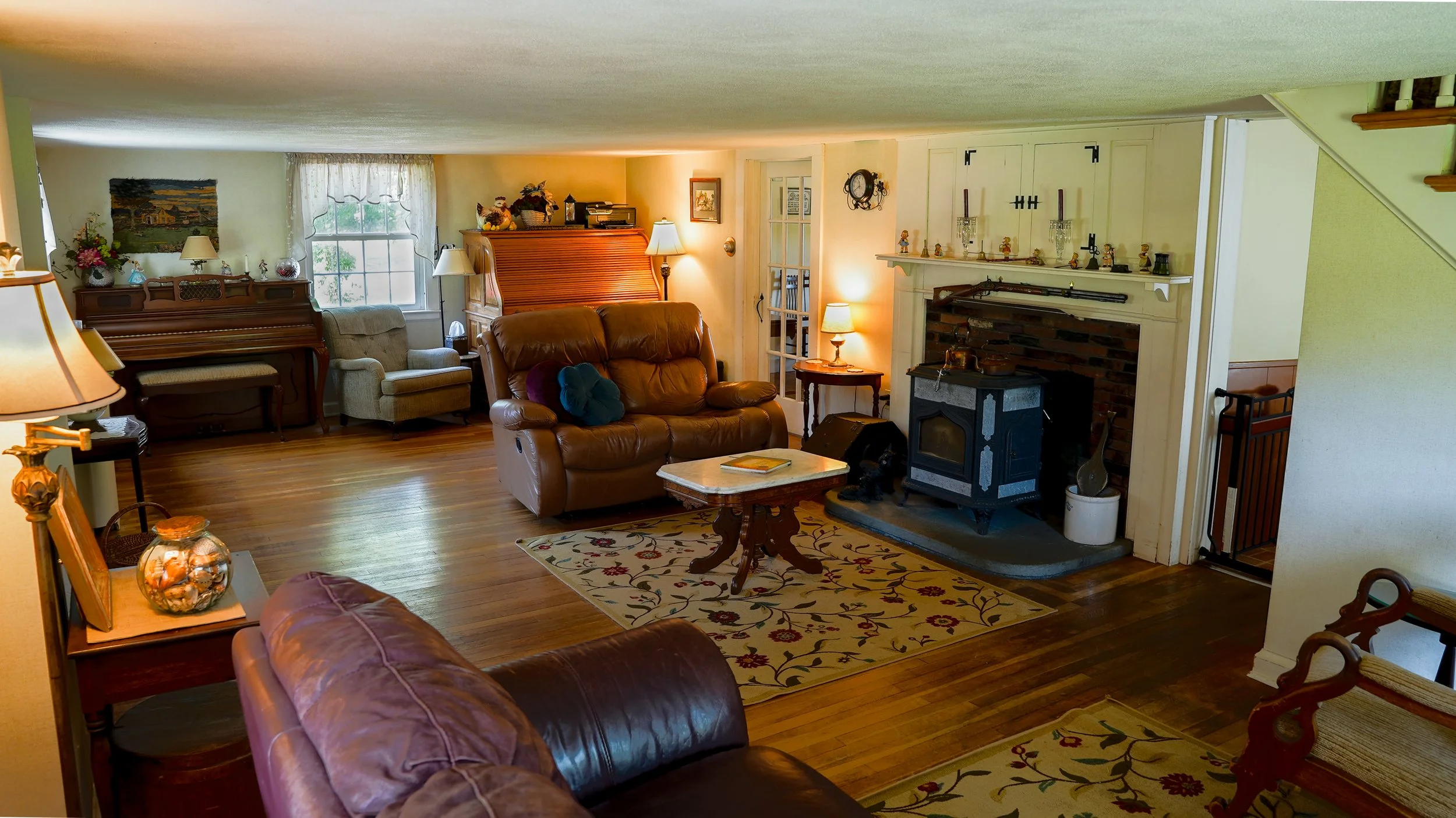 Living room with leather sofas, wooden floor, brick fireplace, piano, various lamps, and home decor.