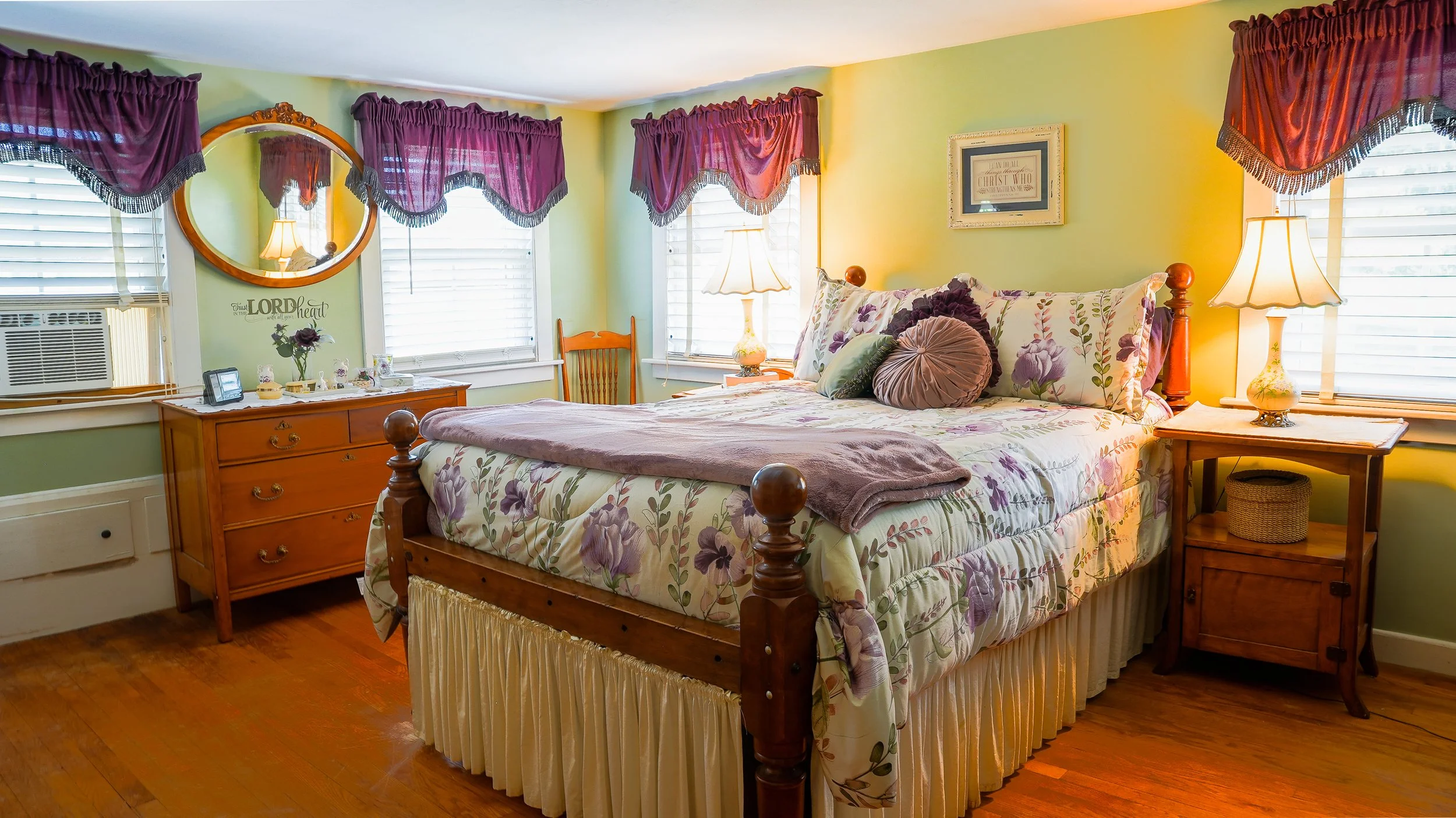 A cozy bedroom with a wooden bed frame, floral bedding, and multiple pillows. There are three windows with purple valances, two bedside lamps, a wooden dresser with decorative items, and a mirror on the wall. The walls are painted light green, and the floor is hardwood.